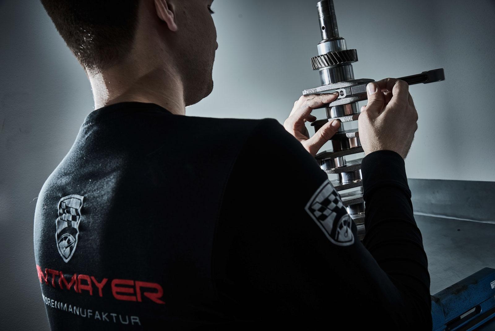 Eine Person, die ein schwarzes Hemd mit einem Logo auf dem Rücken trägt, verwendet einen Messschieber, um ein mechanisches Metallteil in einer Werkstatt zu messen.