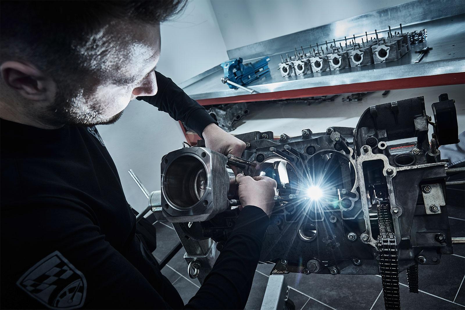 Eine Person arbeitet in einer Werkstatt mit Werkzeugen an einem Motorblock, um dessen Komponenten zusammenzubauen oder zu reparieren, wobei ein helles Licht den Arbeitsbereich beleuchtet. Motorteile und Werkzeuge sind auf einem Tisch im Hintergrund zu sehen.
