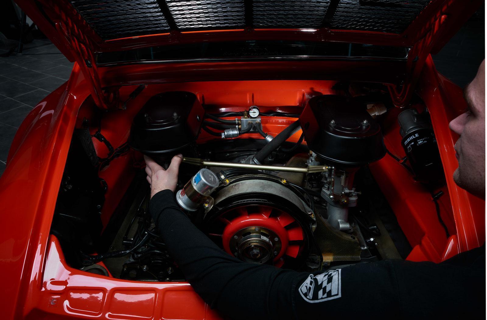 Eine Person arbeitet am Motor eines roten Oldtimers, wobei die Motorabdeckung geöffnet ist und verschiedene mechanische Teile und Komponenten freigelegt werden.