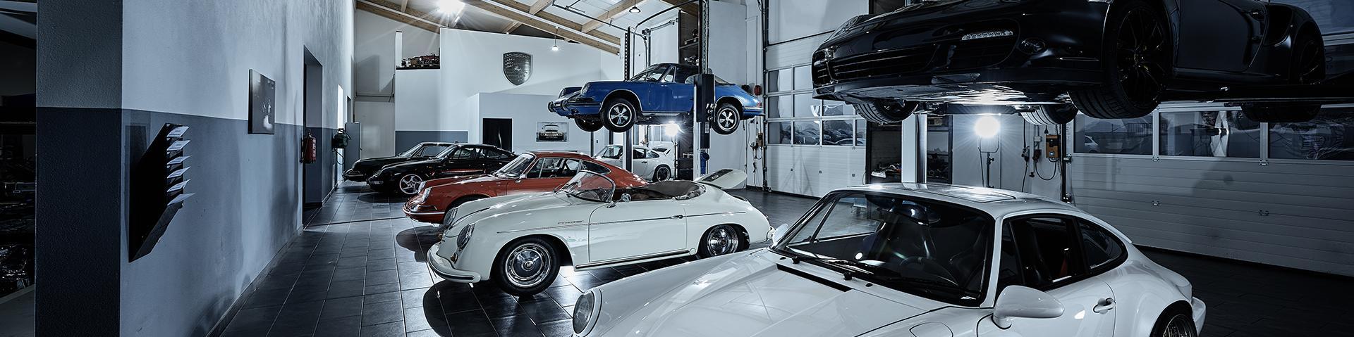 Eine Garage mit mehreren klassischen und modernen Porsche-Fahrzeugen, darunter weiße, rote, blaue und schwarze Modelle, einige auf Hebebühnen, andere auf dem glänzend schwarzen Fliesenboden unter hellen Deckenleuchten geparkt.