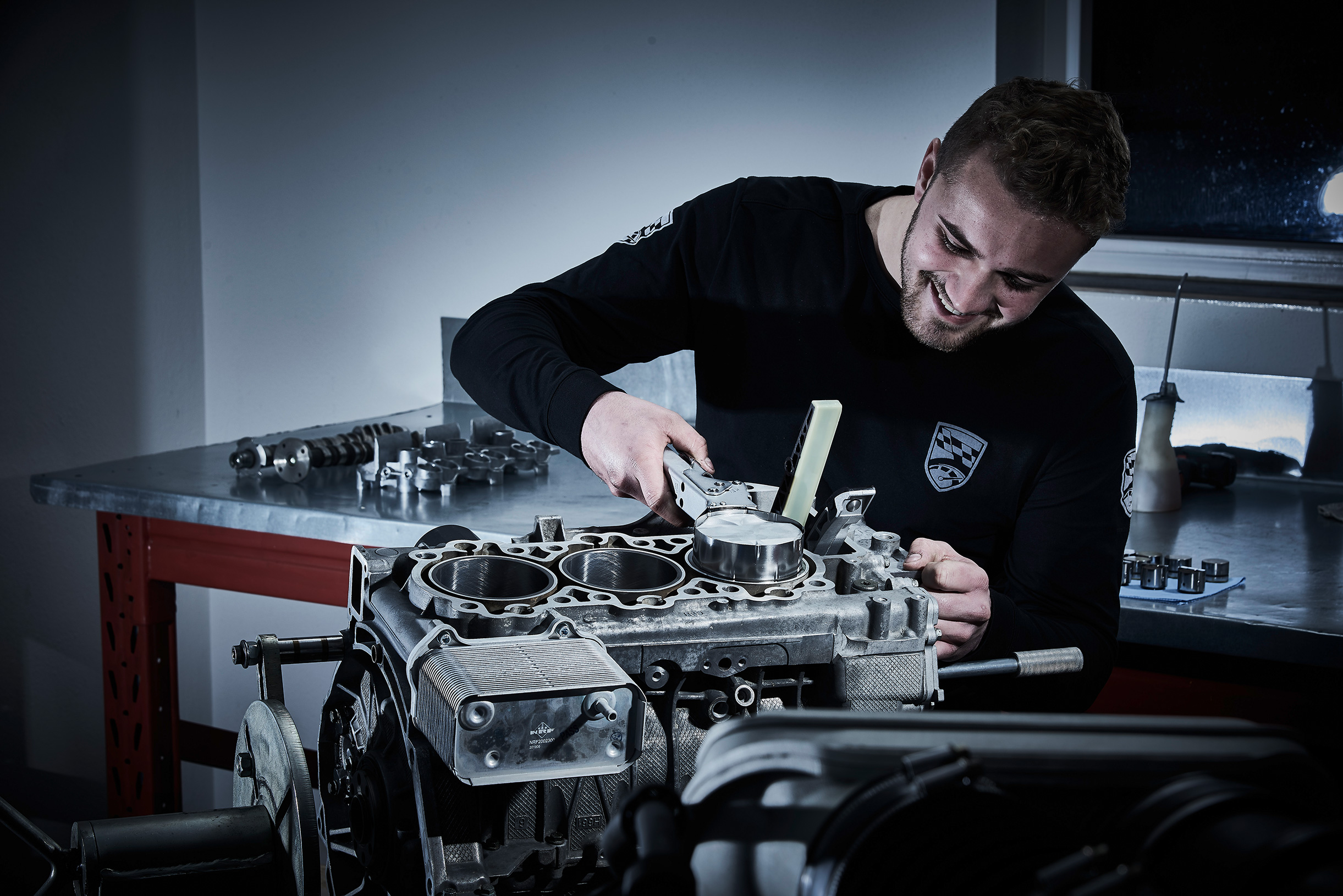 Ein Mann lächelt, während er an einem Automotor arbeitet und ein Werkzeug zum Einstellen von Teilen verwendet. Hinter ihm, in einer gut beleuchteten Werkstatt, liegen Motorteile und Werkzeuge auf der Werkbank.