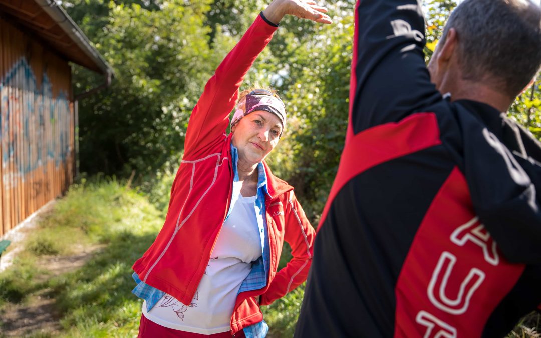 Robert_Treitinger_Trainer©invisions.at-10 Eine ältere Frau und ein älterer Mann in sportlicher Kleidung strecken sich gemeinsam im Freien und heben einen Arm über den Kopf. Sie stehen neben einem Holzgebäude mit Grün und Sonnenlicht im Hintergrund.