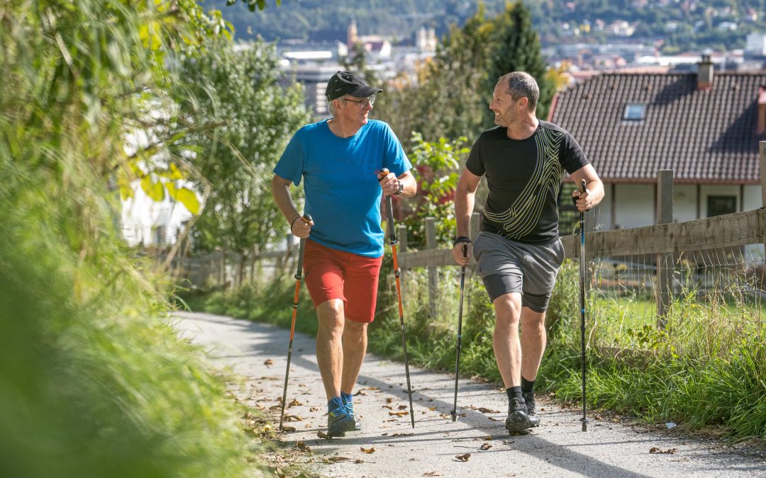 Robert_Treitinger_Trainer©invisions.at-2 Zwei Männer gehen mit Stöcken auf einem gepflasterten Weg im Freien spazieren, tragen Sportkleidung, unterhalten sich und lächeln sich an, im Hintergrund sind Häuser und Grünanlagen zu sehen.
