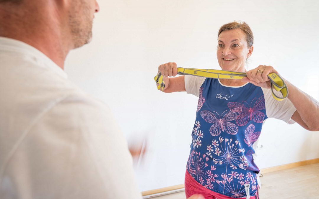 Robert_Treitinger_Trainer©invisions.at-21 Eine ältere Frau lächelt, während sie in einem Haus ein gelbes Widerstandsband vor der Brust hält. Im Vordergrund steht ein Mann in einem weißen Hemd, der sie anleitet oder ihr hilft.