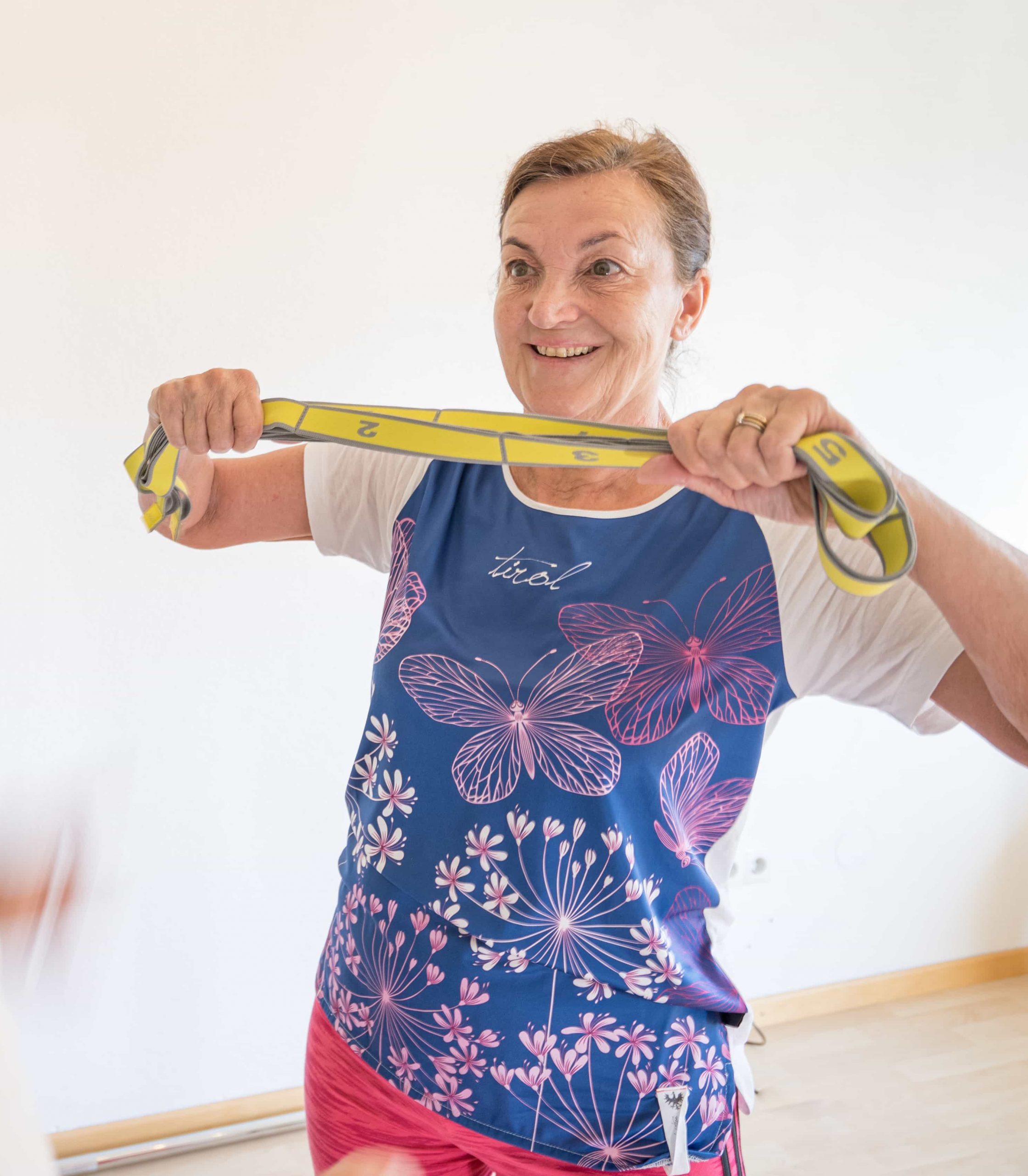 Eine lächelnde ältere Frau in einem Hemd mit Schmetterlingsmuster trainiert in einer Halle und zieht mit beiden Händen an einem gelben Widerstandsband.