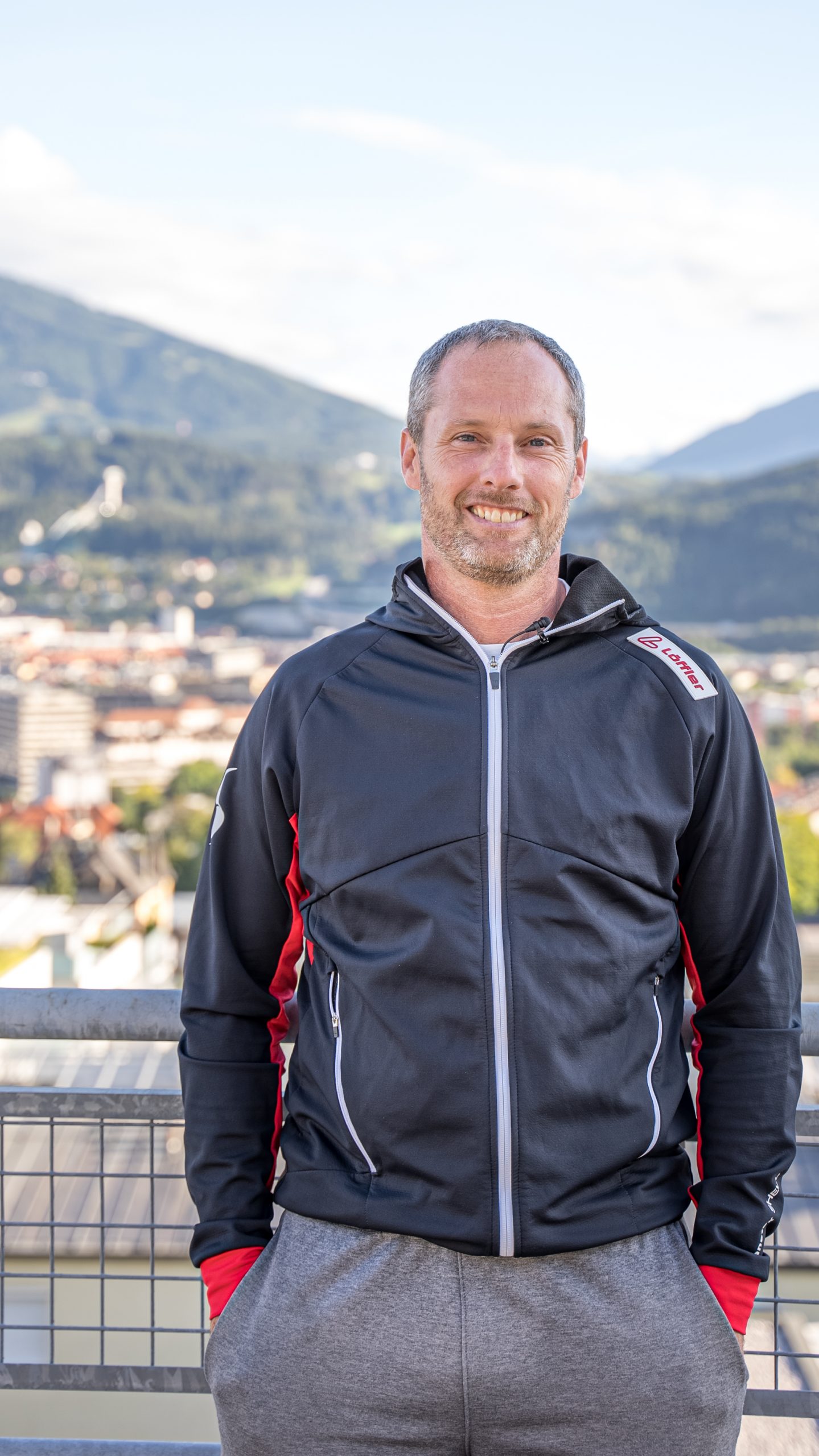 Ein Mann in einer schwarz-roten Jacke steht im Freien und lächelt, während im Hintergrund eine verschwommene Stadtlandschaft und grüne Berge unter einem teilweise bewölkten Himmel zu sehen sind.