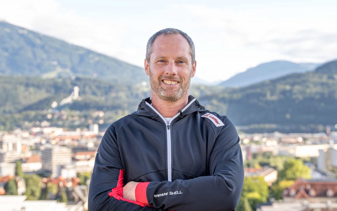 Robert_Treitinger_Trainer©invisions.at-30 Ein Mann in einer schwarzen Jacke steht im Freien mit verschränkten Armen, lächelnd, vor dem Hintergrund einer Stadt und grüner Berge unter einem teilweise bewölkten Himmel.