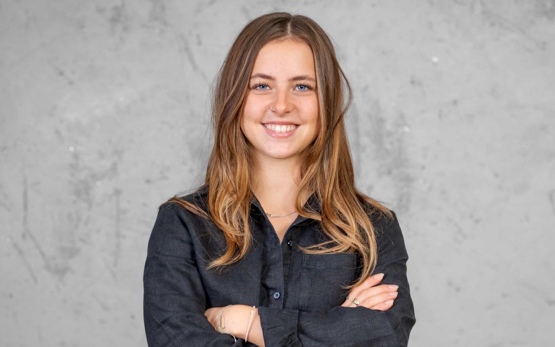 Eine junge Frau mit langem hellbraunem Haar und schwarzem Hemd steht mit verschränkten Armen und lächelt in die Kamera vor einem schlichten grauen Hintergrund.