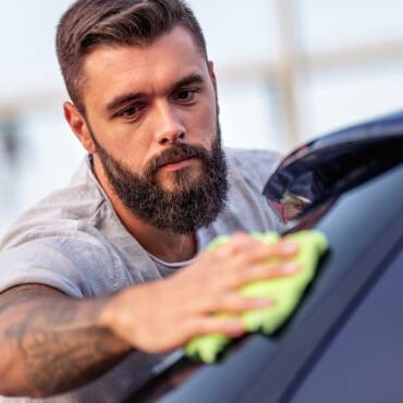 Ein bärtiger Mann reinigt mit einem grünen Lappen die Außenseite eines Autos und konzentriert sich dabei auf seine Aufgabe.