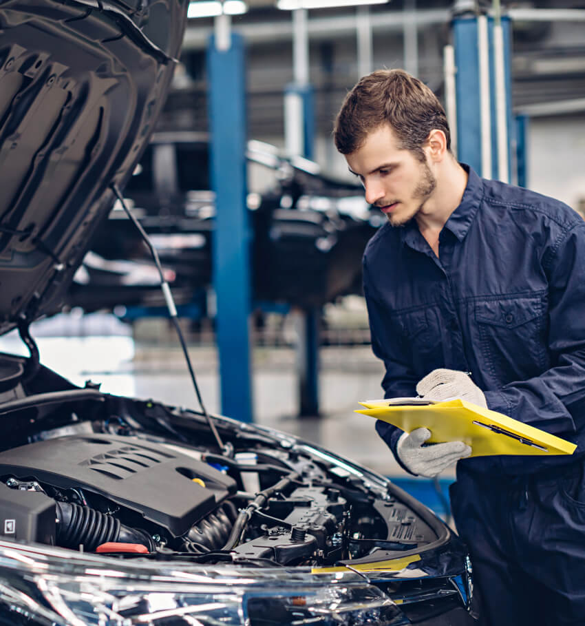 Ein Mechaniker in einem blauen Overall und mit Handschuhen inspiziert einen Automotor bei geöffneter Motorhaube, mit einem gelben Klemmbrett und einem Stift in der Hand, in einer Autowerkstatt mit Hebebühnen im Hintergrund.