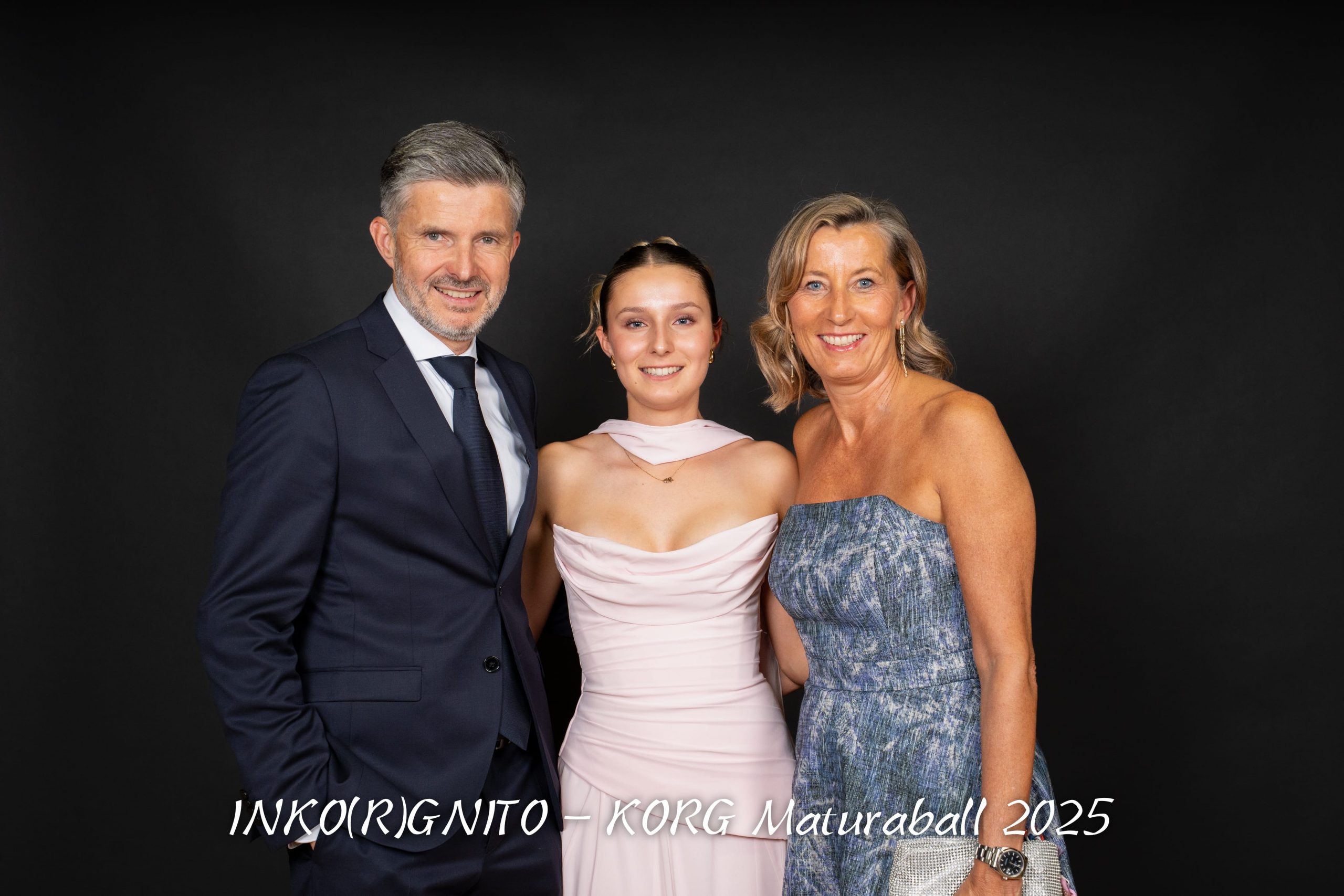 Drei Personen, zwei Erwachsene und eine junge Frau in formeller Kleidung, stehen lächelnd zusammen vor einem schwarzen Hintergrund. Der Text am unteren Rand lautet: INKO(R)GNITO - KORG Maturaball 2025.