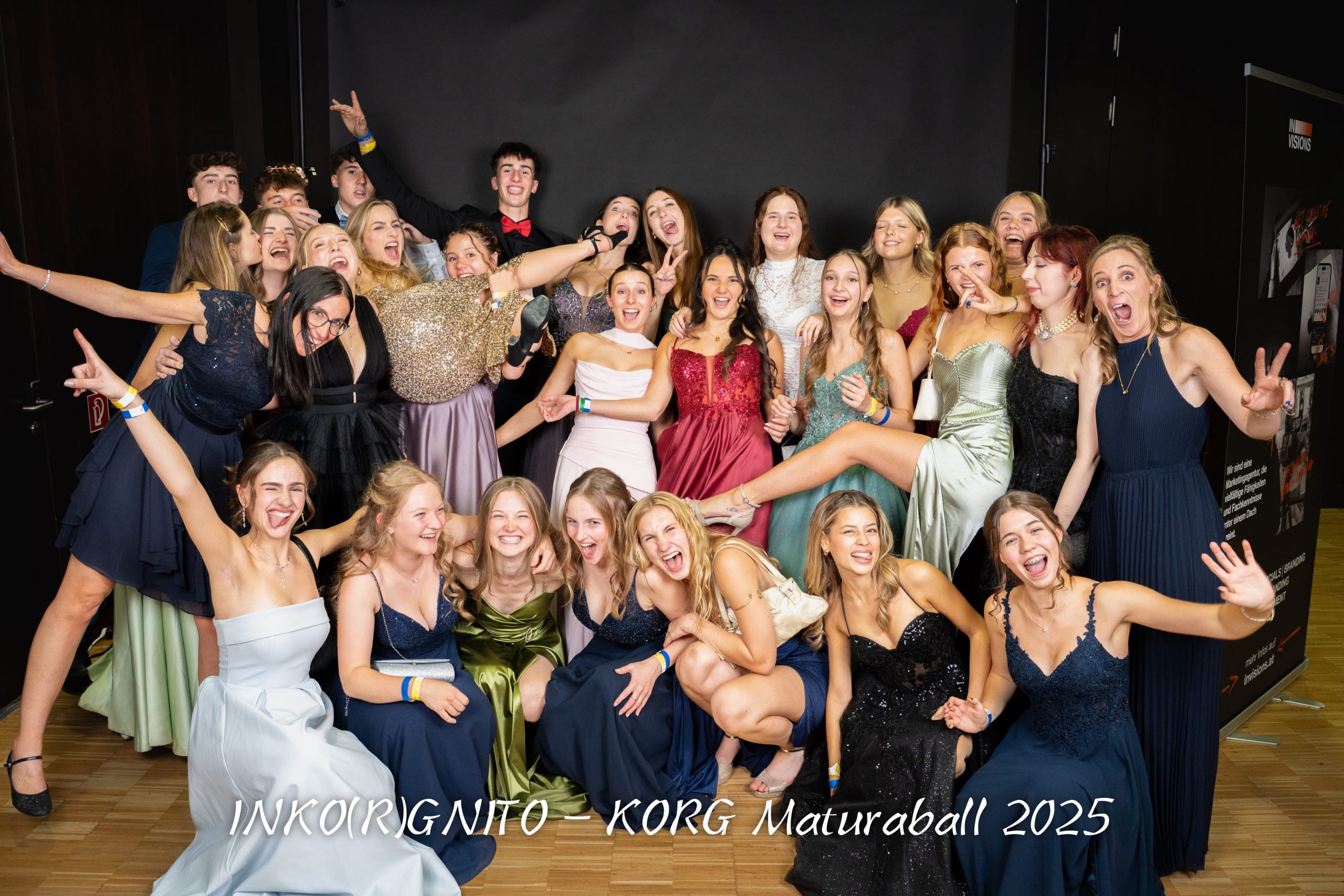 Eine große Gruppe junger Leute in formeller Kleidung posiert lächelnd und lachend auf dem KORG Maturaball 2025. Sie feiern auf einem Holzboden mit einem schwarzen Hintergrund hinter ihnen.