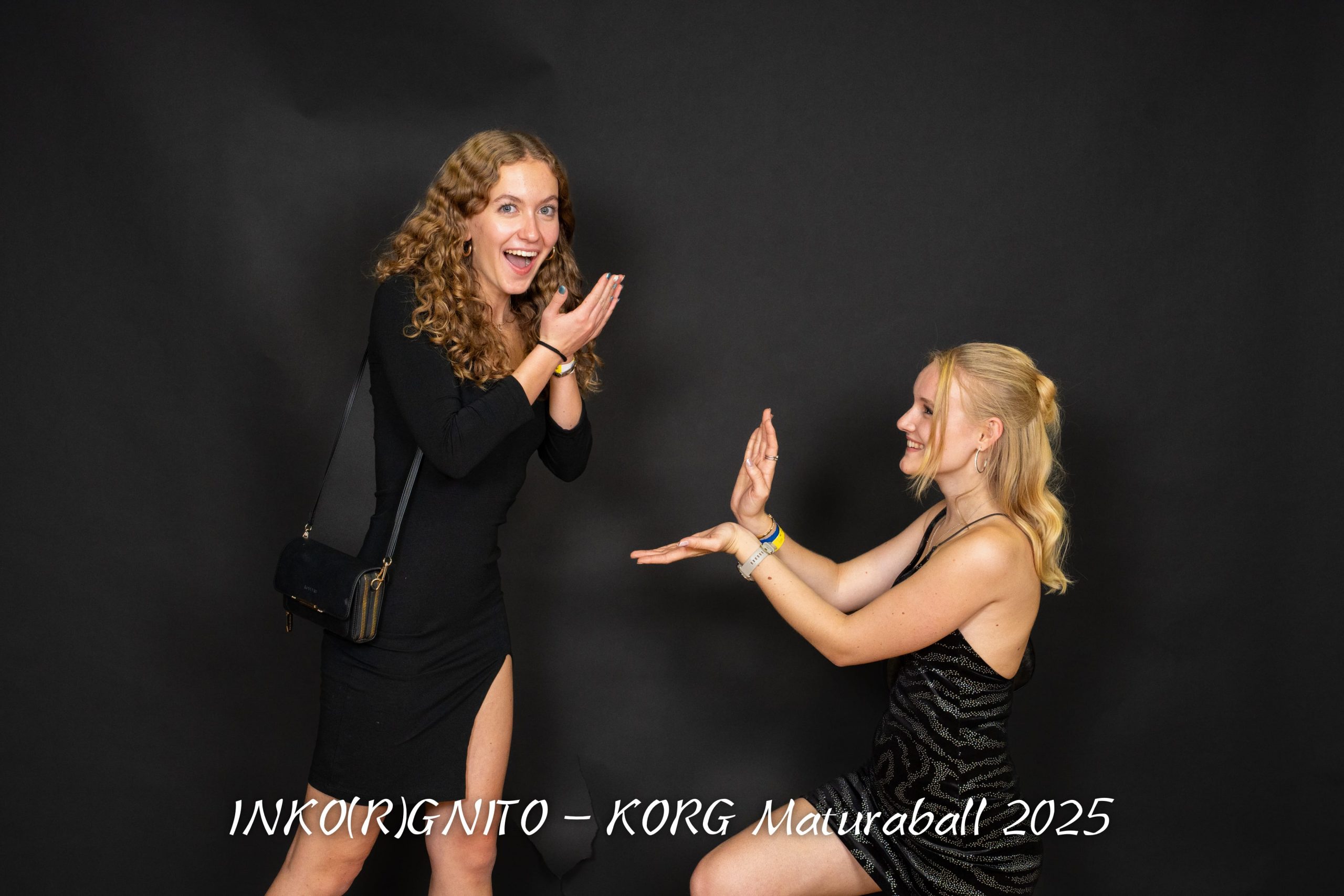 Zwei junge Frauen posieren vor einem schwarzen Hintergrund; eine steht und hält sich überrascht die Hände vors Gesicht, während die andere kniet und einen Antrag mimt. Die Bildunterschrift lautet "INKOIRGINITO - KORG Maturaball 2025".