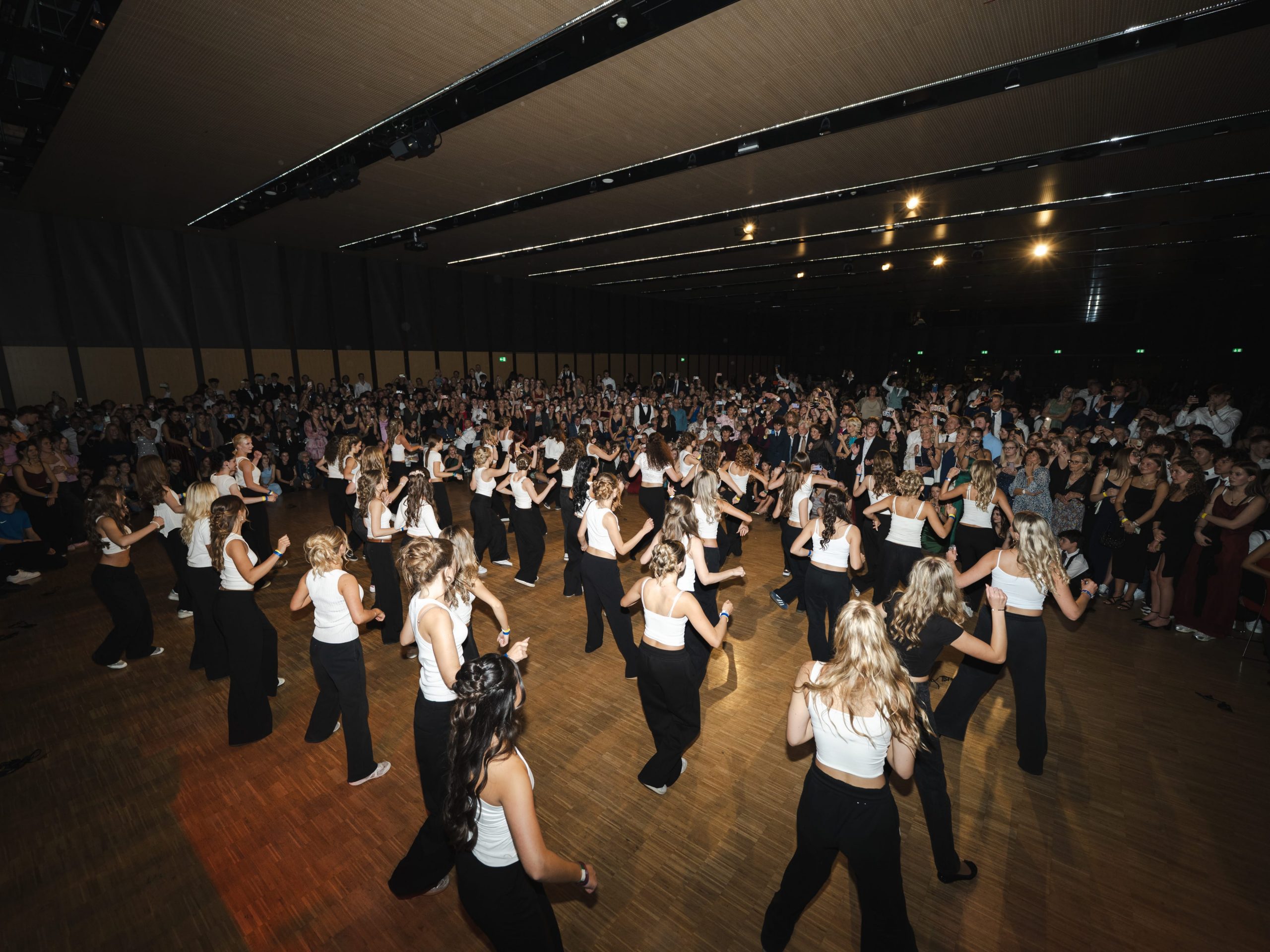Eine große Gruppe von Frauen in schwarzen Hosen und weißen Oberteilen führt in der Mitte einer großen Halle einen Synchrontanz auf, umgeben von einer Menge aufmerksamer Zuschauer.