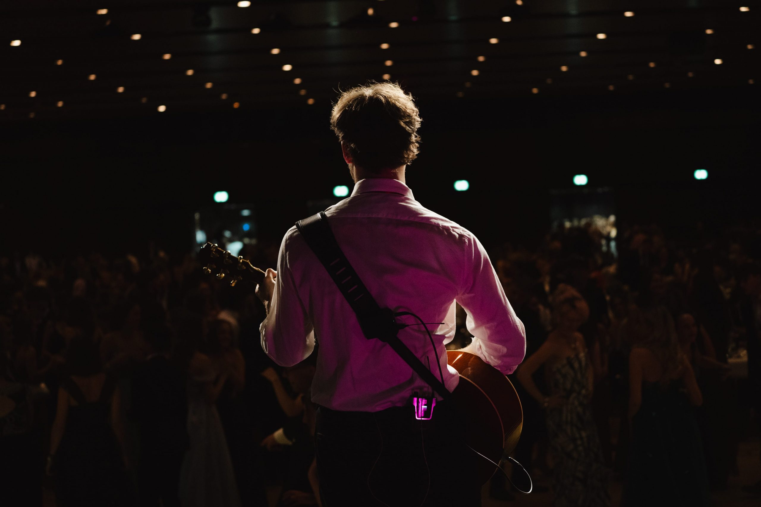 Ein Gitarrist in einem weißen Hemd steht auf der Bühne und blickt auf ein verschwommenes Publikum in einem schwach beleuchteten Saal, das von Bühnenscheinwerfern angestrahlt wird, und bereitet sich auf seinen Auftritt vor.