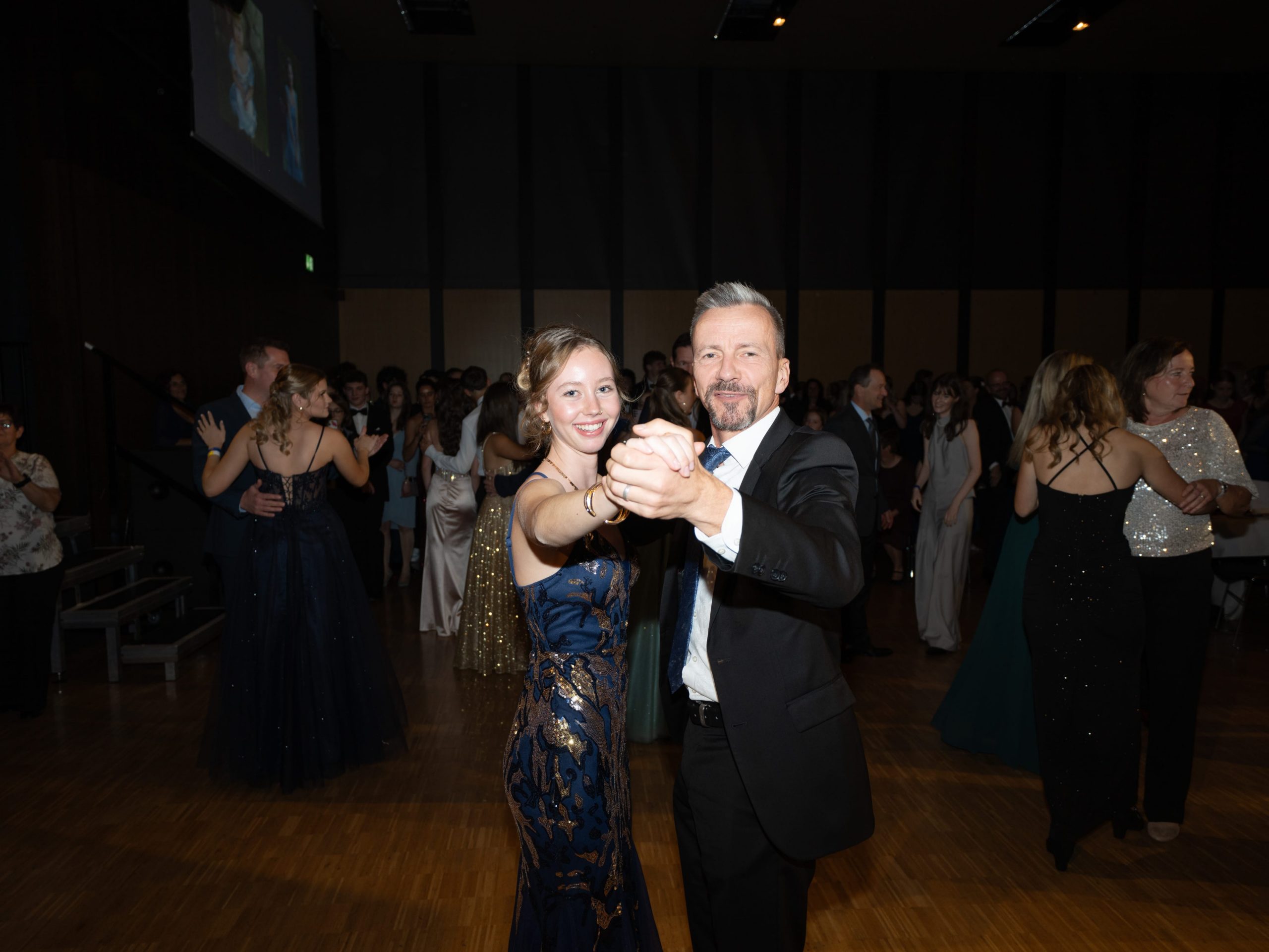 Ein lächelnder Mann und eine lächelnde Frau in formeller Kleidung tanzen zusammen auf einer Veranstaltung im Ballsaal, umgeben von anderen elegant gekleideten Paaren auf einer hölzernen Tanzfläche.