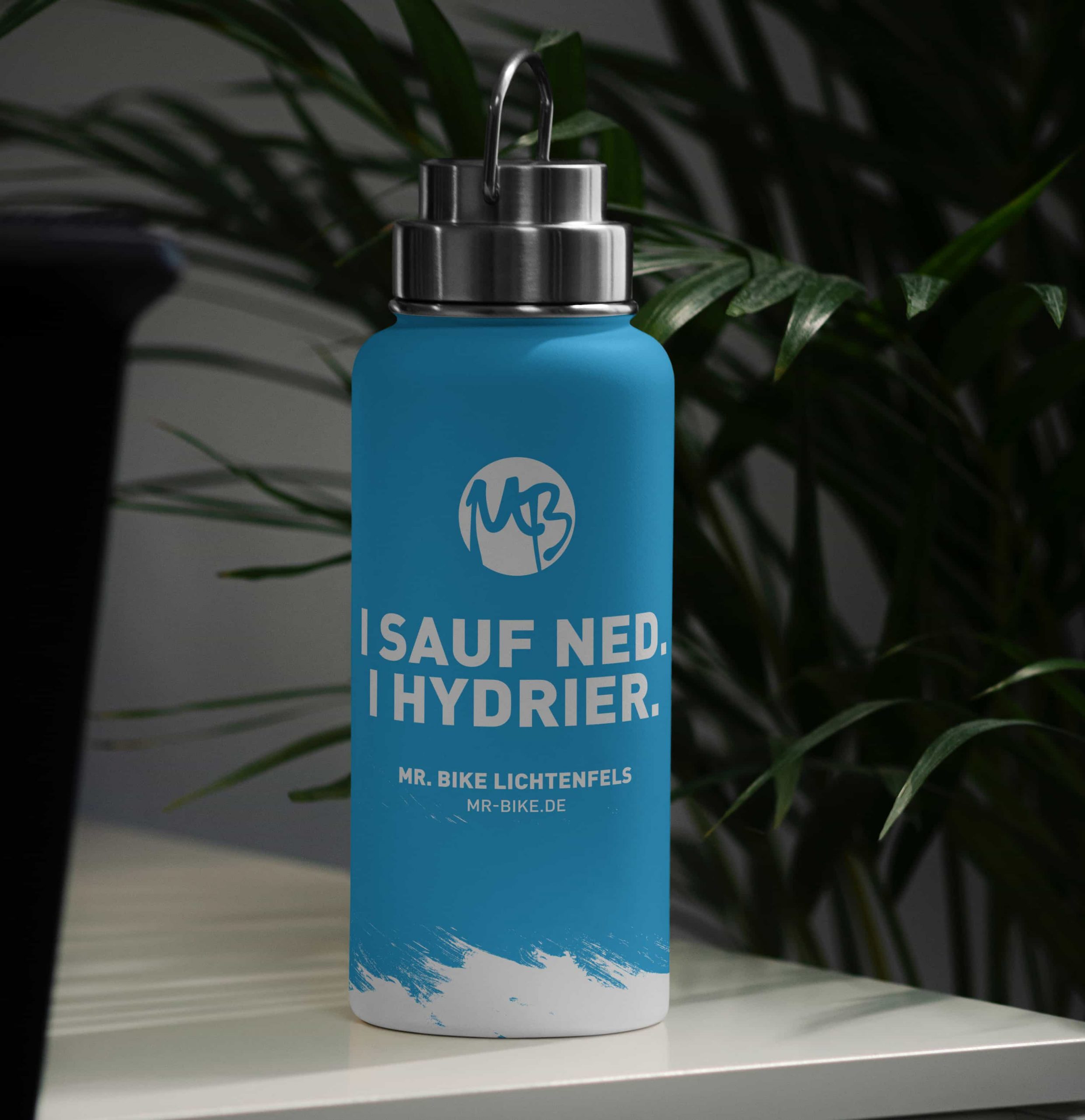 Eine blaue wiederverwendbare Wasserflasche mit silbernem Verschluss und Griff steht auf einem Tisch. Auf der Flasche steht in weißer Schrift: "I SAUF NED. I HYDRIER." und das Branding von MR. BIKE LICHTENFELS. Im Hintergrund ist eine Pflanze zu sehen.