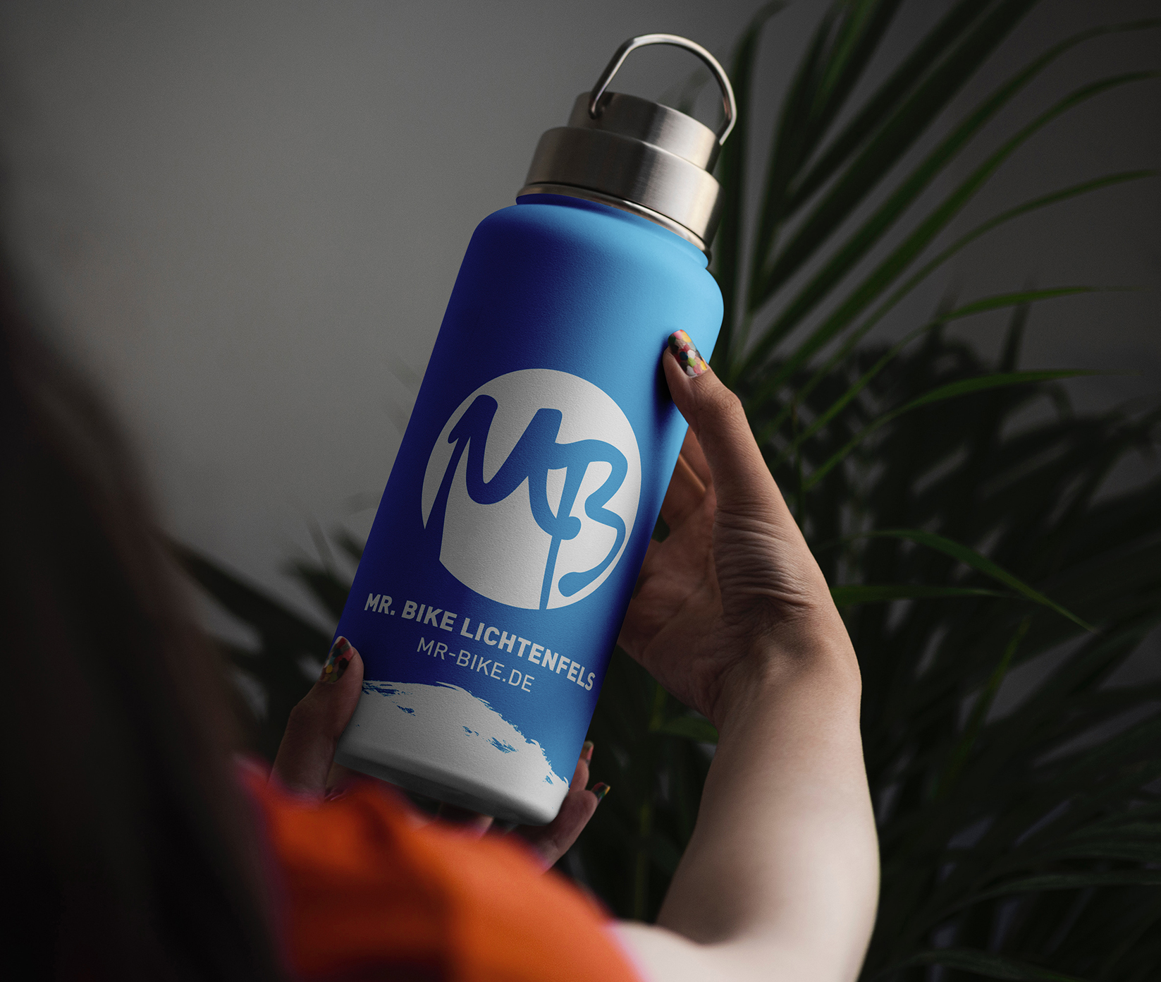 Eine Person hält eine blaue Wasserflasche mit einem silbernen Deckel, auf dem das Logo MB und der Text MR. BIKE LICHTENFELS und MR-BIKE.DE in Weiß. Im Hintergrund sind grüne Blätter zu sehen.