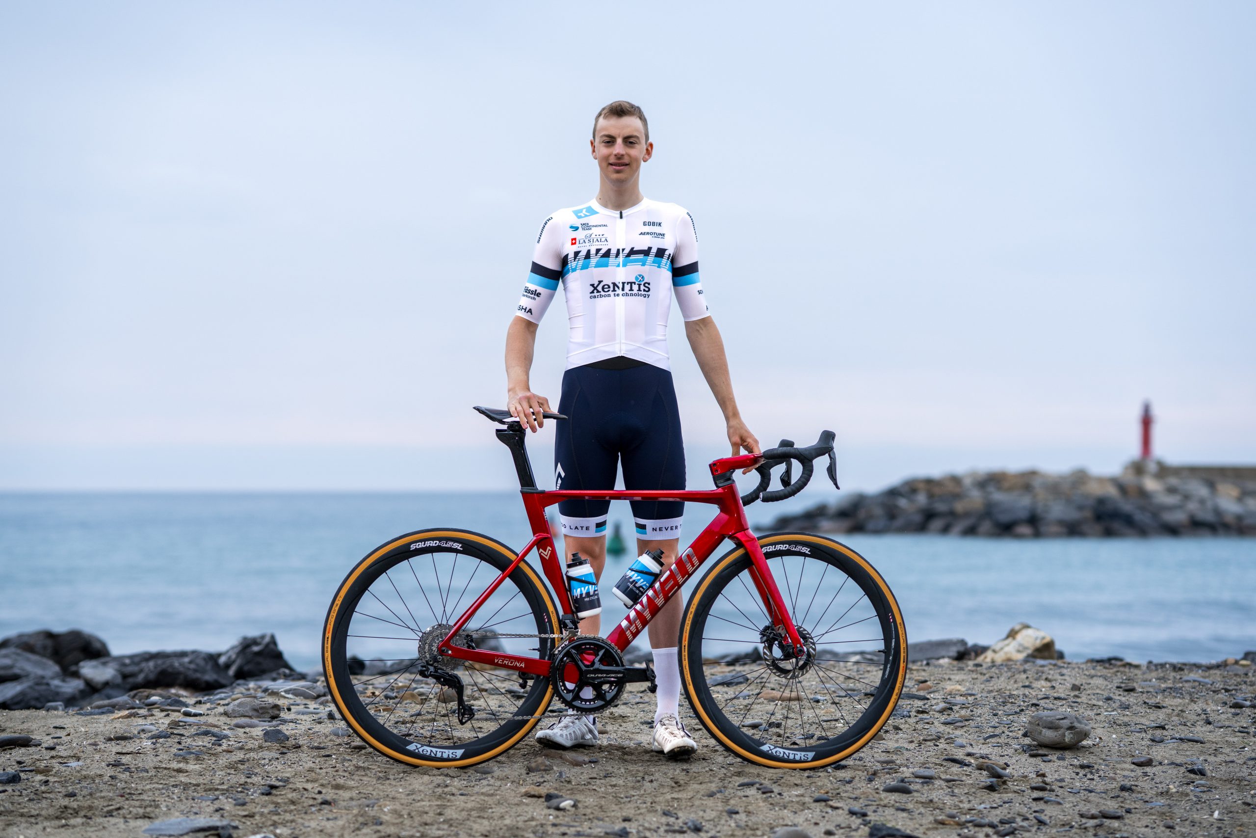 Ein Radfahrer in einem weiß-blauen Rennanzug steht an einem felsigen Strand, in der Hand ein rotes Rennrad, im Hintergrund das Meer und ein entfernter roter Leuchtturm.