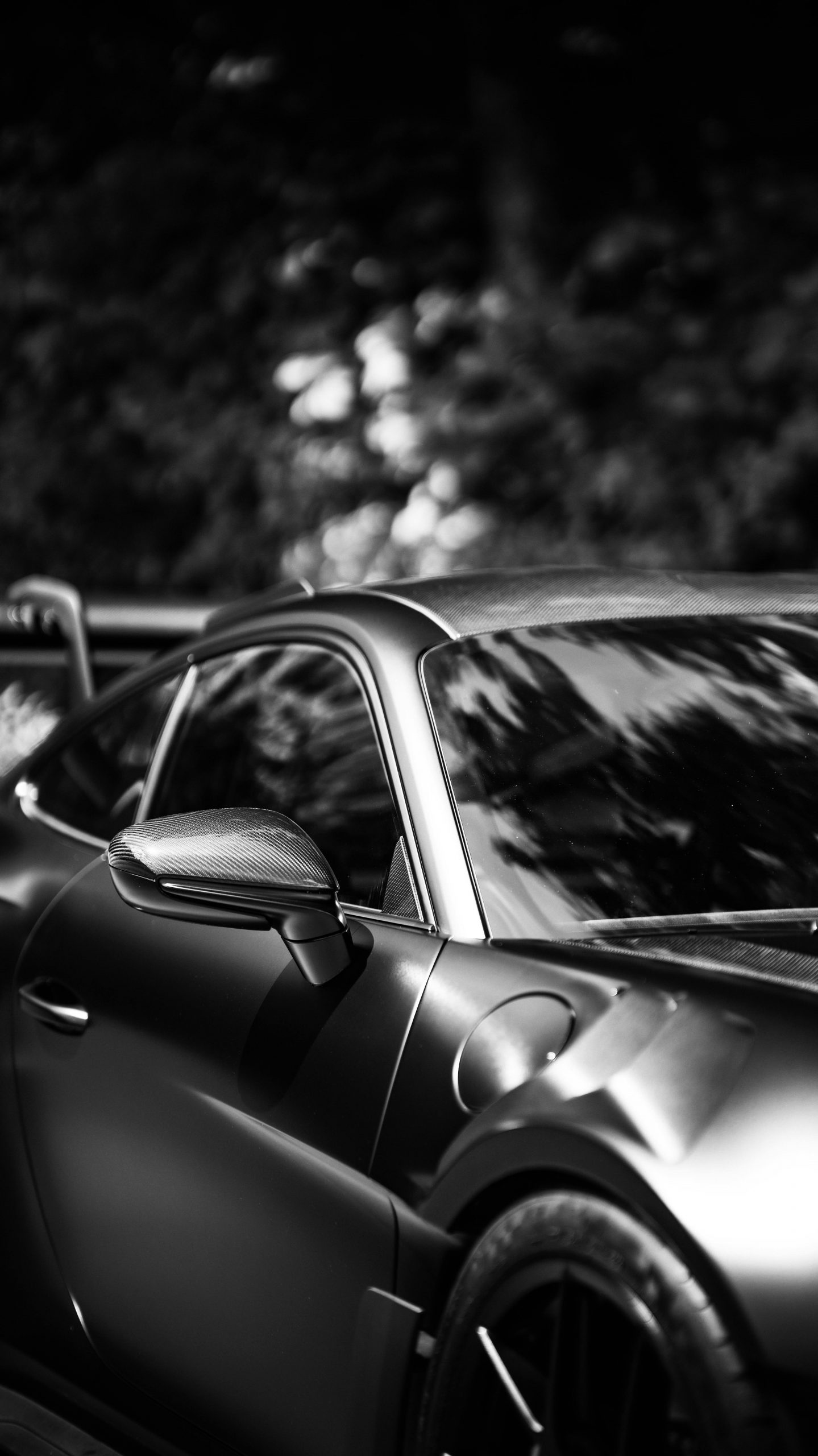 Schwarz-Weiß-Nahaufnahme eines schnittigen Sportwagens, die sich auf den Seitenspiegel, das Fenster, die Tür und einen Teil des Heckflügels konzentriert, mit unscharfen Bäumen, die sich auf der Oberfläche des Wagens spiegeln.