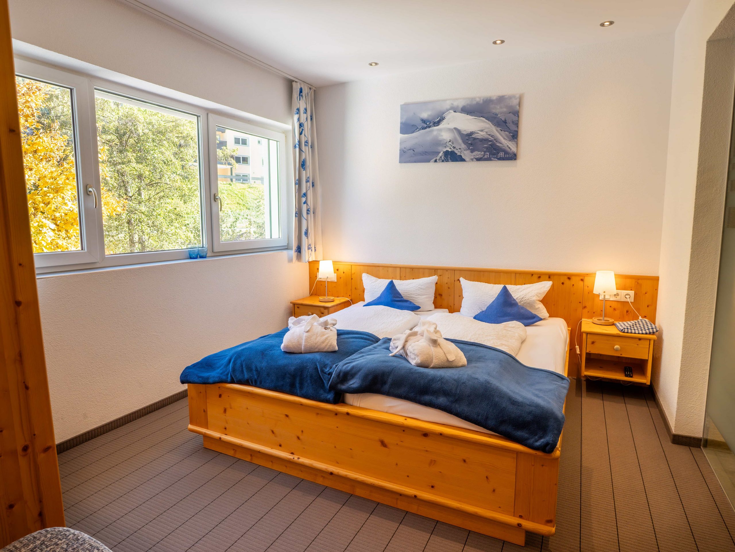 Ein gemütliches Hotelzimmer mit einem Doppelbett, blau-weißem Bettzeug, zwei Handtuchschwänen, Holzmöbeln, Nachttischlampen, einem Fenster mit Blick auf Bäume und einem verschneiten Bergbild an der Wand.