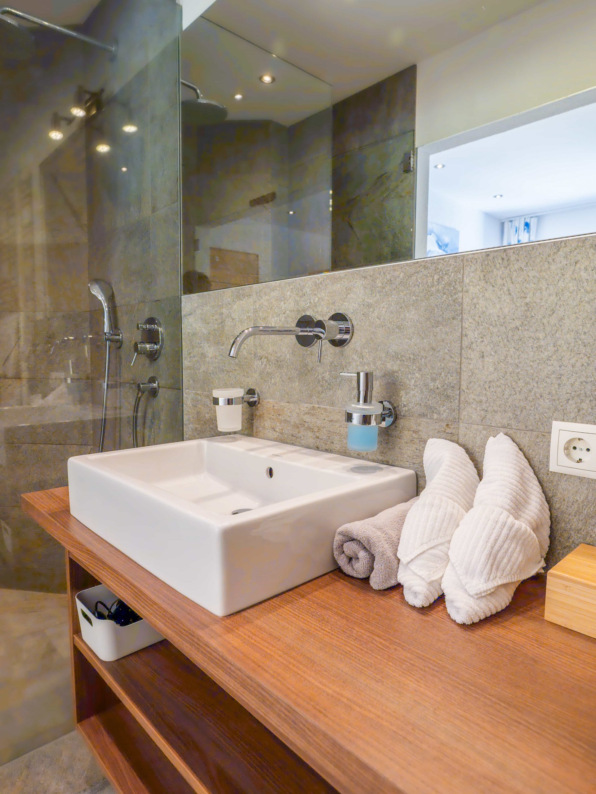 Modernes Badezimmer mit einem quadratischen weißen Waschbecken auf einer hölzernen Arbeitsplatte, wandmontierten Armaturen, Toilettenartikeln und drei ordentlich gerollten weißen Handtüchern. Der Duschbereich ist mit Glasplatten und grauen Steinfliesen ausgestattet.