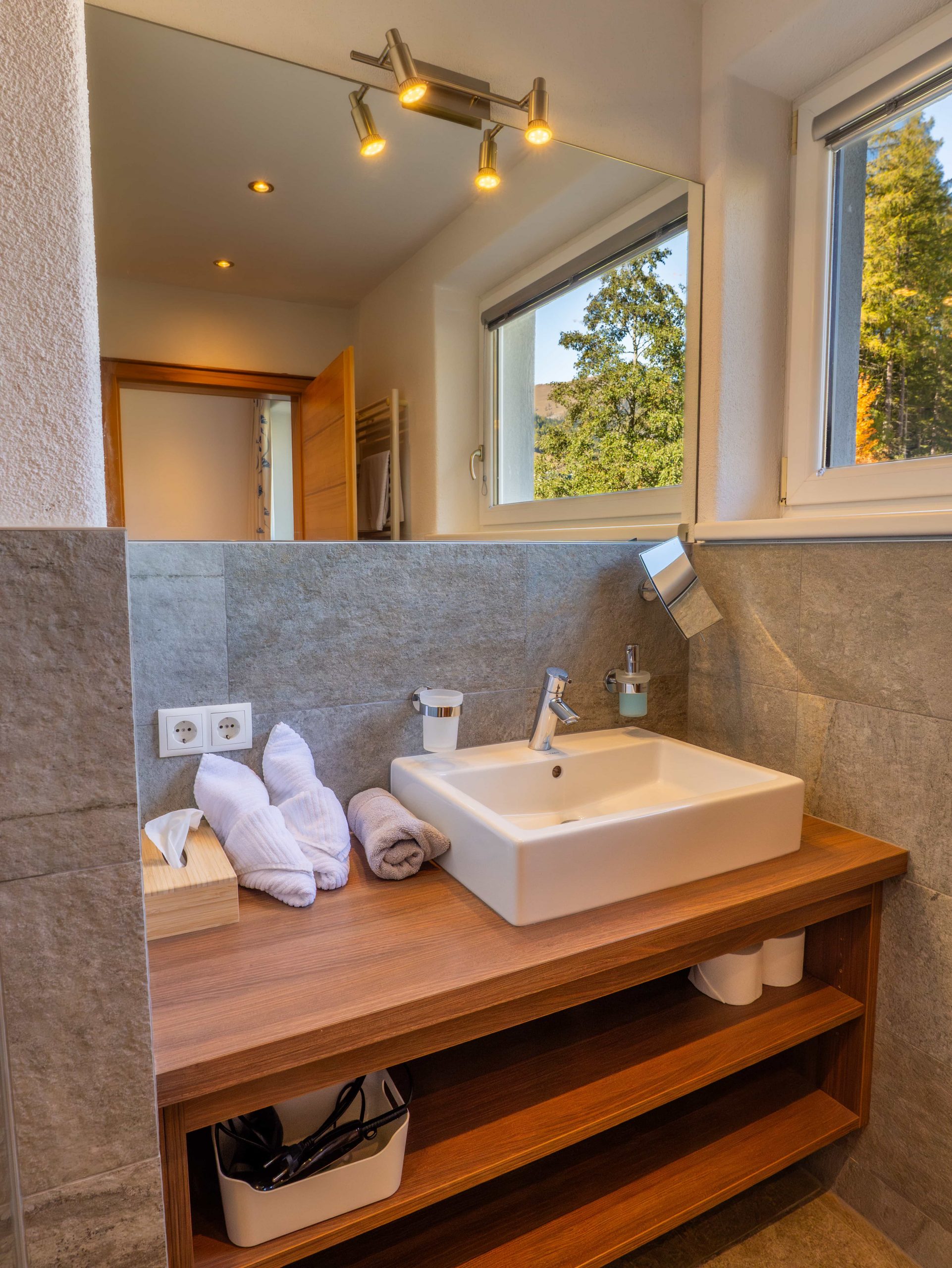 Modernes Badezimmer mit einem quadratischen weißen Waschbecken auf einer hölzernen Arbeitsplatte, ordentlich aufgerollten Handtüchern, Toilettenartikeln, einem Spiegel und großen Fenstern mit Blick auf grüne Bäume draußen. Unter dem Waschbecken sind grau geflieste Wände und Regale mit Körben zu sehen.