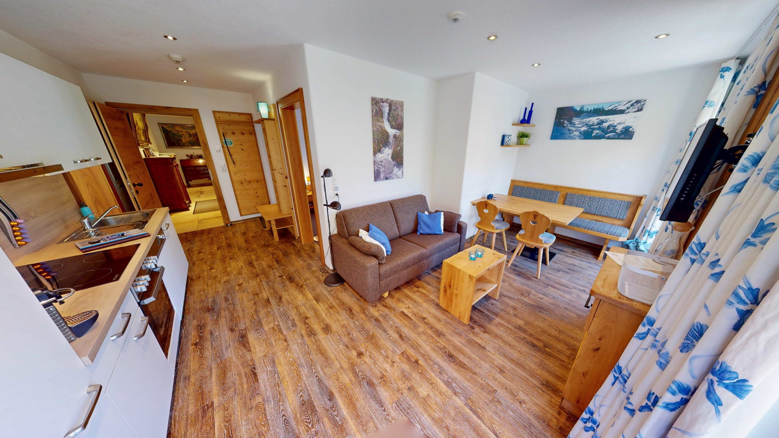 Ein gemütlicher, moderner Wohnraum mit Holzböden, einer kleinen Küche auf der linken Seite, einem braunen Sofa, einem hölzernen Esstisch mit gepolsterter Bank und großen Fenstern mit geblümten Vorhängen, die natürliches Licht hereinlassen.