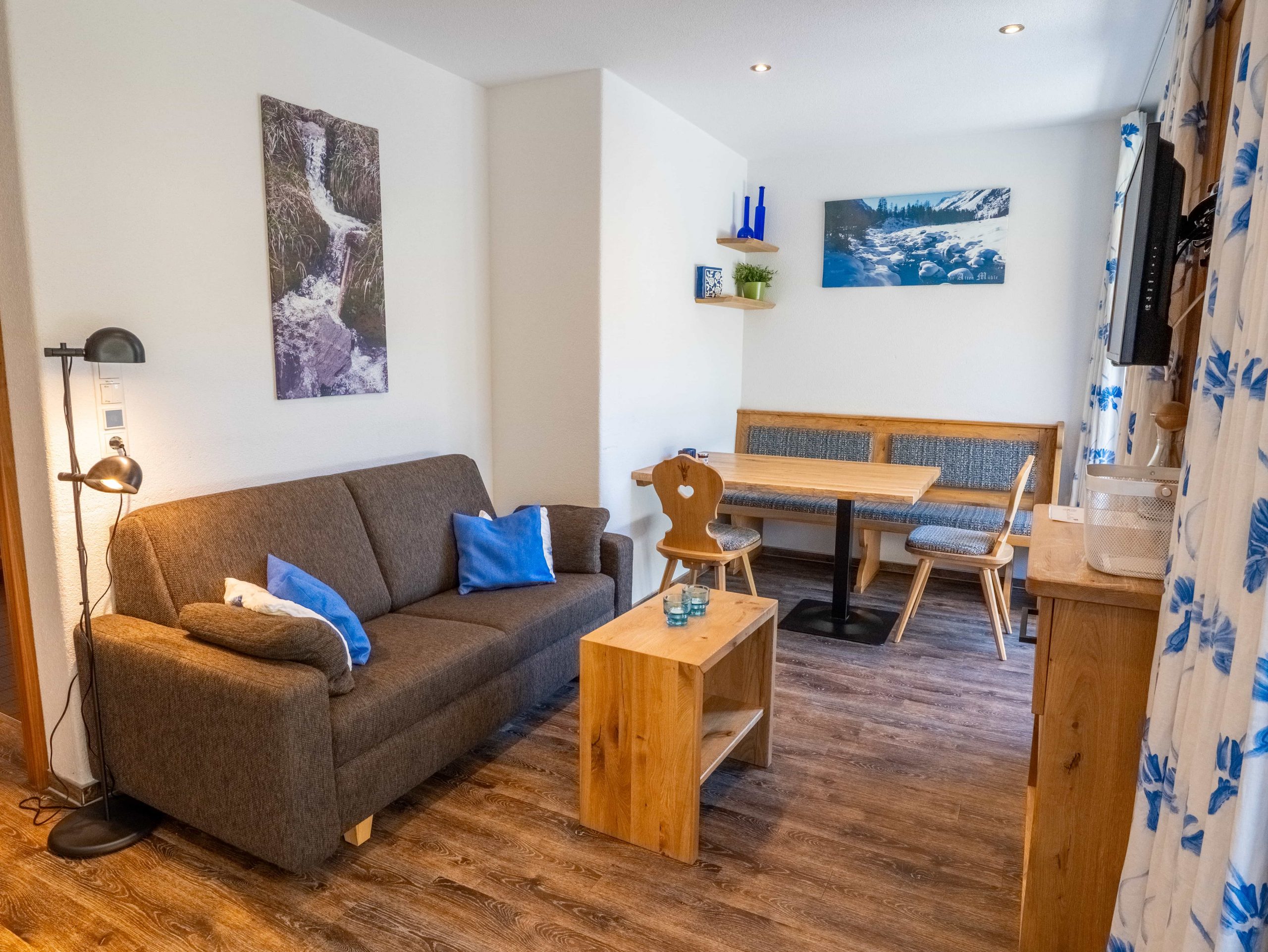 Gemütliches Wohnzimmer mit einem grauen Sofa, blauen Kissen, einem Couchtisch aus Holz, einer Essecke mit Bank und Stühlen, Wandkunst, Zimmerpflanzen und gemusterten Vorhängen auf einem Holzboden.