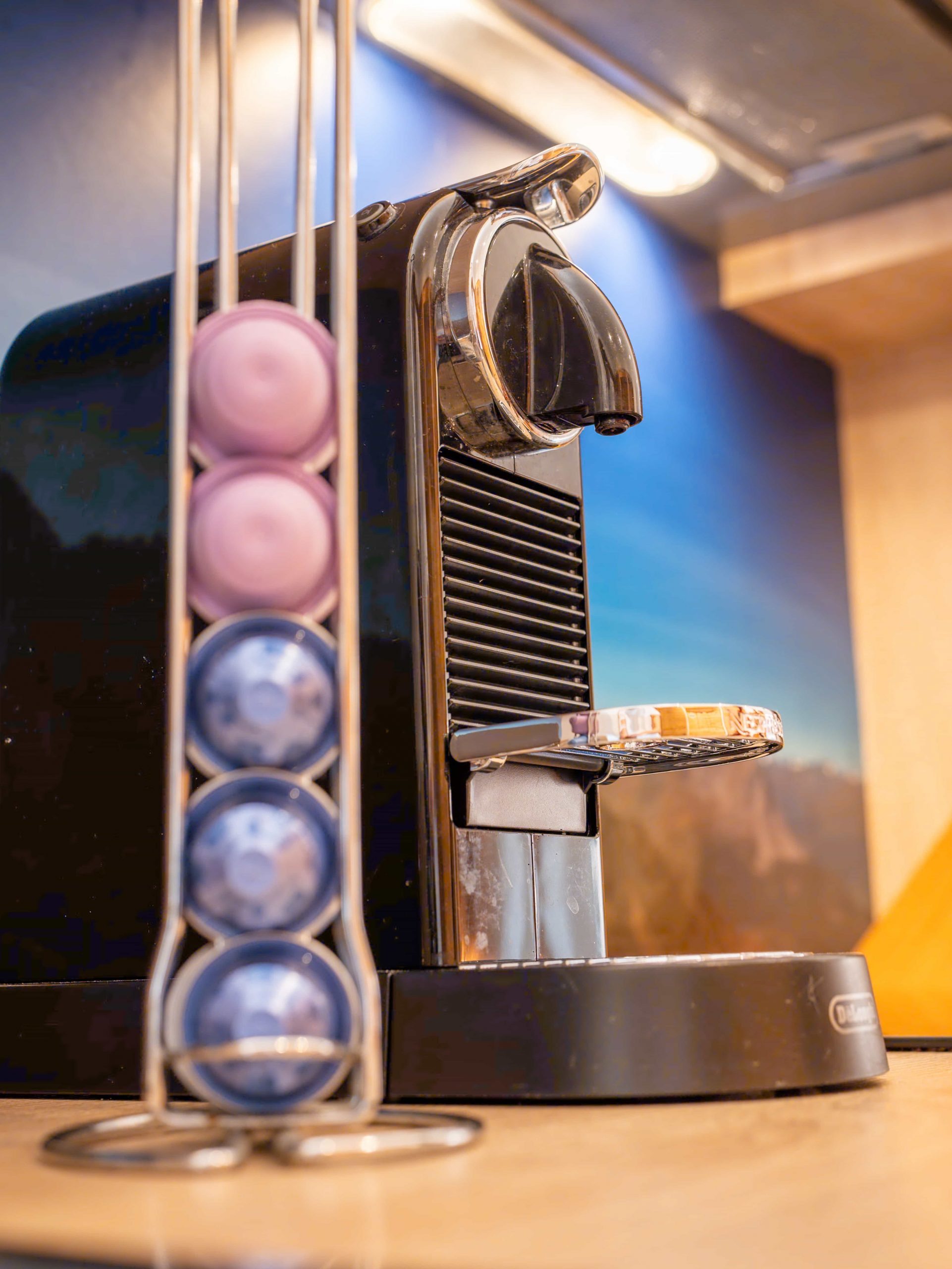 Eine schwarze Espressomaschine steht auf einer Küchentheke neben einem vertikalen Metallbehälter, der Kaffeepads in Lila-, Rosa- und Blautönen enthält. Der Hintergrund zeigt eine Küchenwand und eine Unterschrankbeleuchtung.