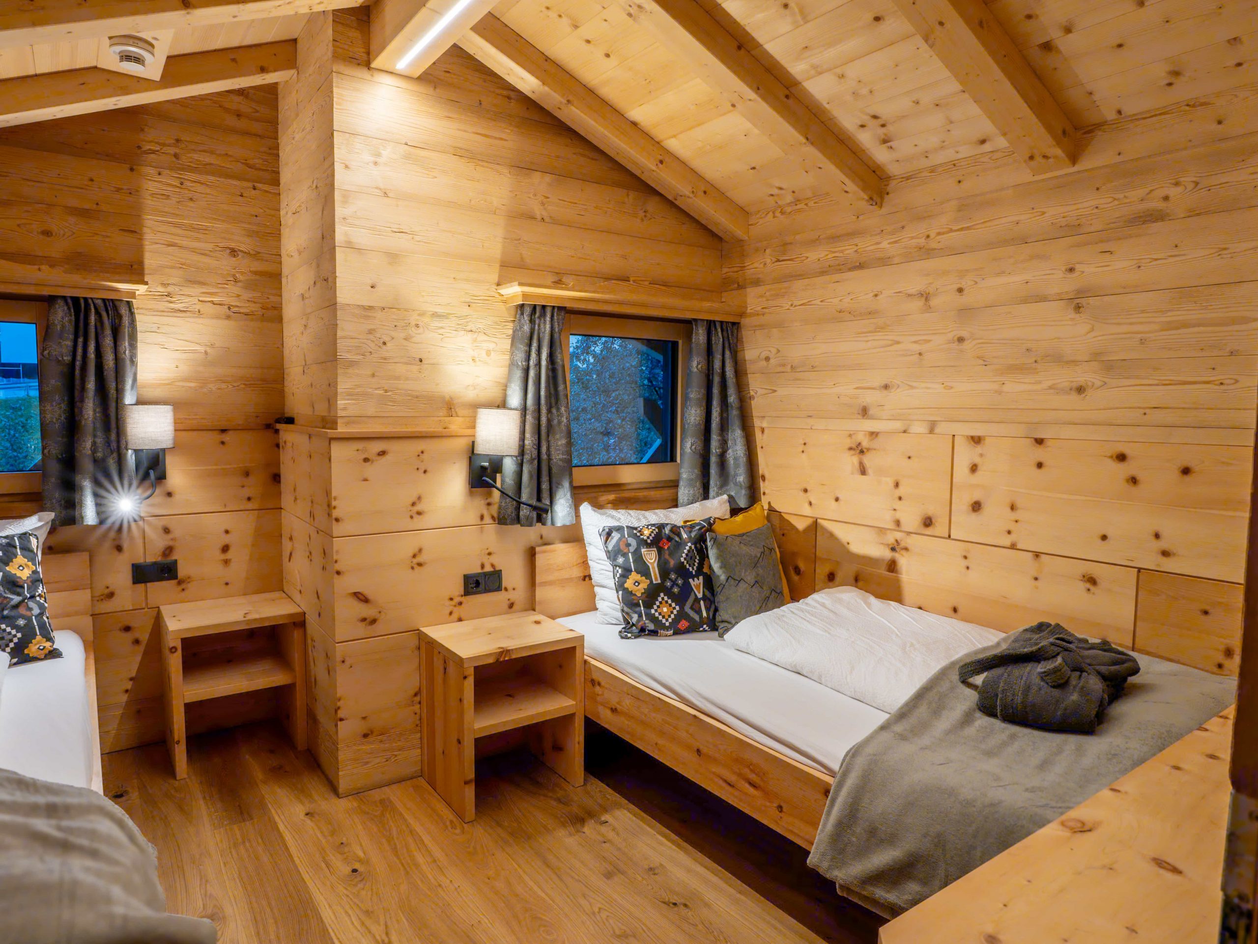 Gemütliches Holzhüttenzimmer mit zwei Einzelbetten, gemusterten Kissen, grauen Decken, Nachttischen, Lampen und großen Fenstern mit Vorhängen. Die warme Umgebungsbeleuchtung unterstreicht die einladende Atmosphäre.