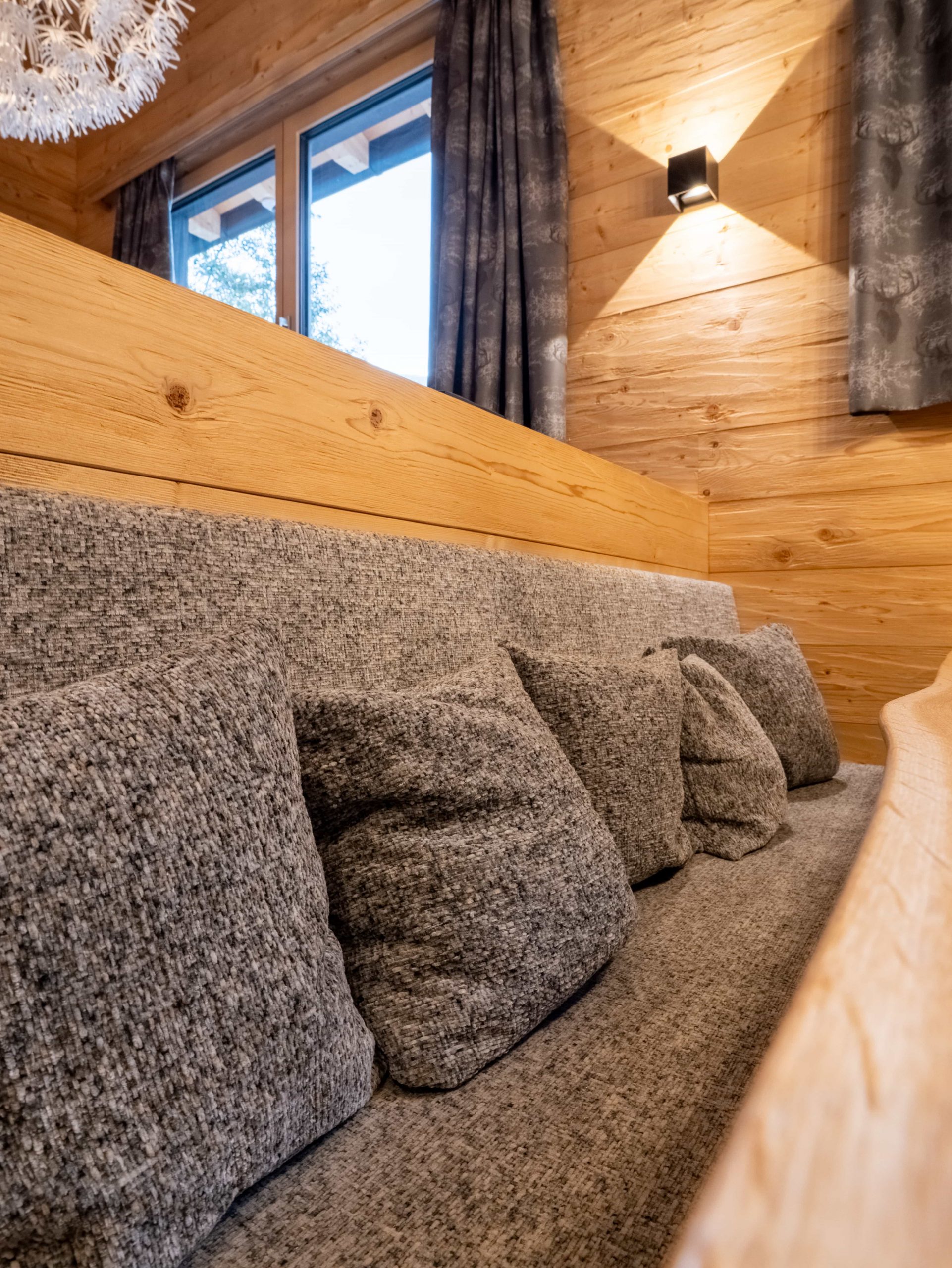 In einem Raum mit holzgetäfelten Wänden, einem Fenster mit dunklen Vorhängen und einer Wandleuchte steht eine gemütliche graue Couch mit mehreren passenden Kissen an einer Holzwand.