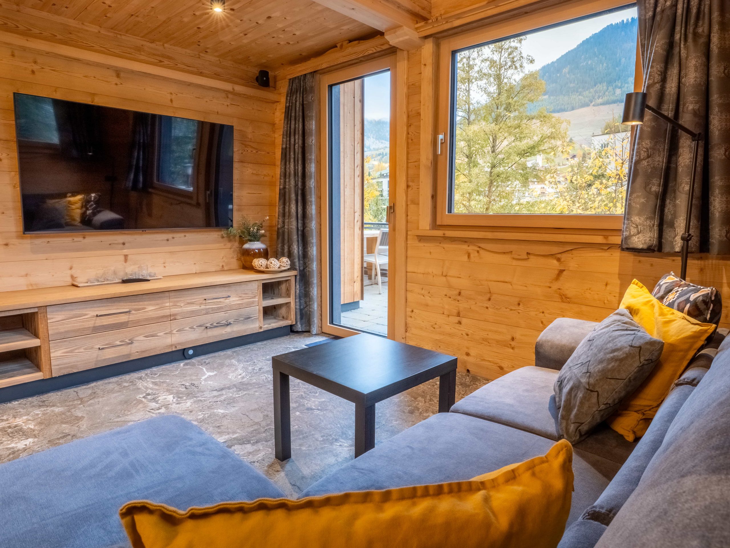 Ein gemütliches Wohnzimmer mit Holzwänden, einem großen Fenster mit Blick auf die Berge, einem grauen Sofa mit gelben Kissen, einem schwarzen Couchtisch, einem Flachbildfernseher und einer Medienkonsole aus Holz.