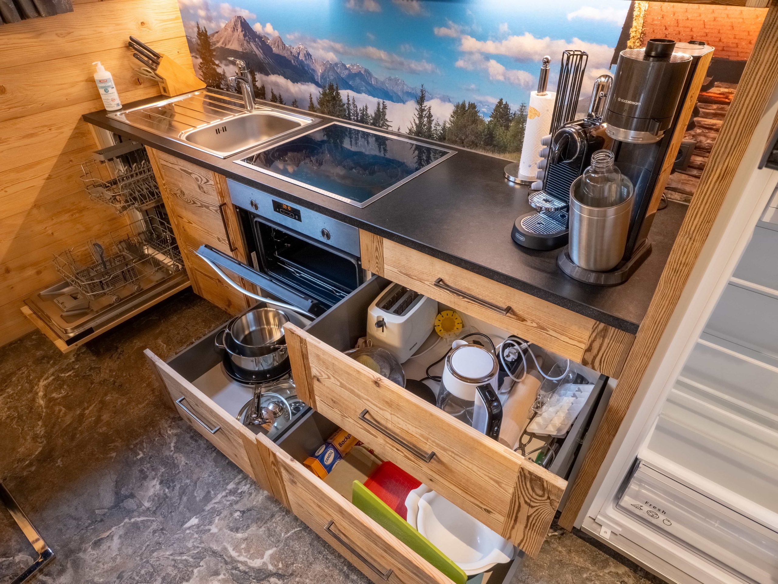 Eine moderne Küche mit offenen Schubladen, in denen Utensilien, Töpfe, ein Toaster und andere Geräte untergebracht sind. Es gibt eine Spüle, eine Herdplatte und eine Aufkantung mit einer malerischen Berglandschaft. Auf der Arbeitsplatte befinden sich eine Kaffeemaschine und andere Küchenutensilien.