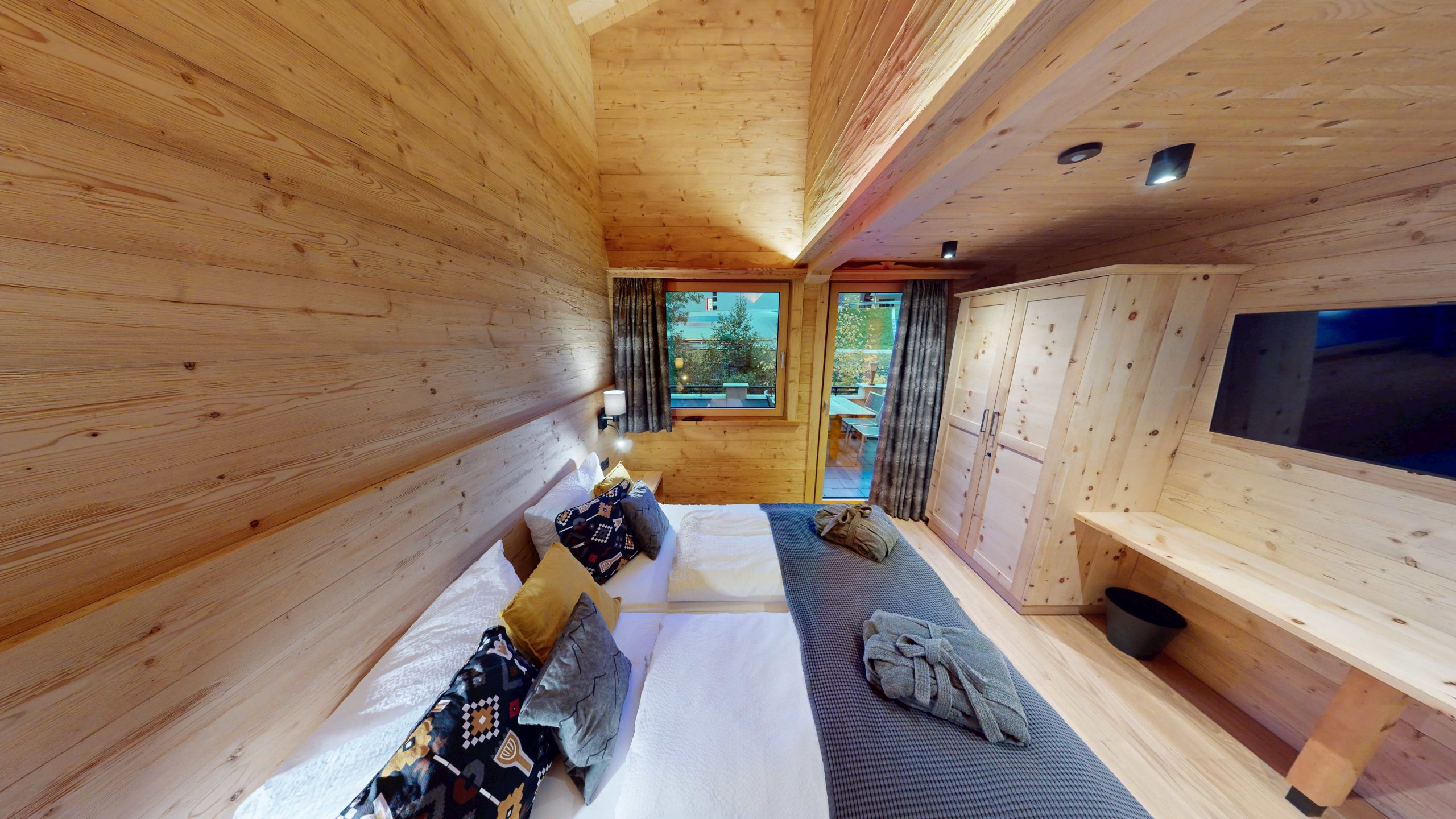 Ein gemütliches Holzschlafzimmer mit einem Doppelbett, bunten Kissen, Nachttischlampen, einem Kleiderschrank, einem an der Wand montierten Fernseher und großen Fenstern mit Blick auf die Bäume draußen. Die warme Beleuchtung unterstreicht die einladende Atmosphäre.