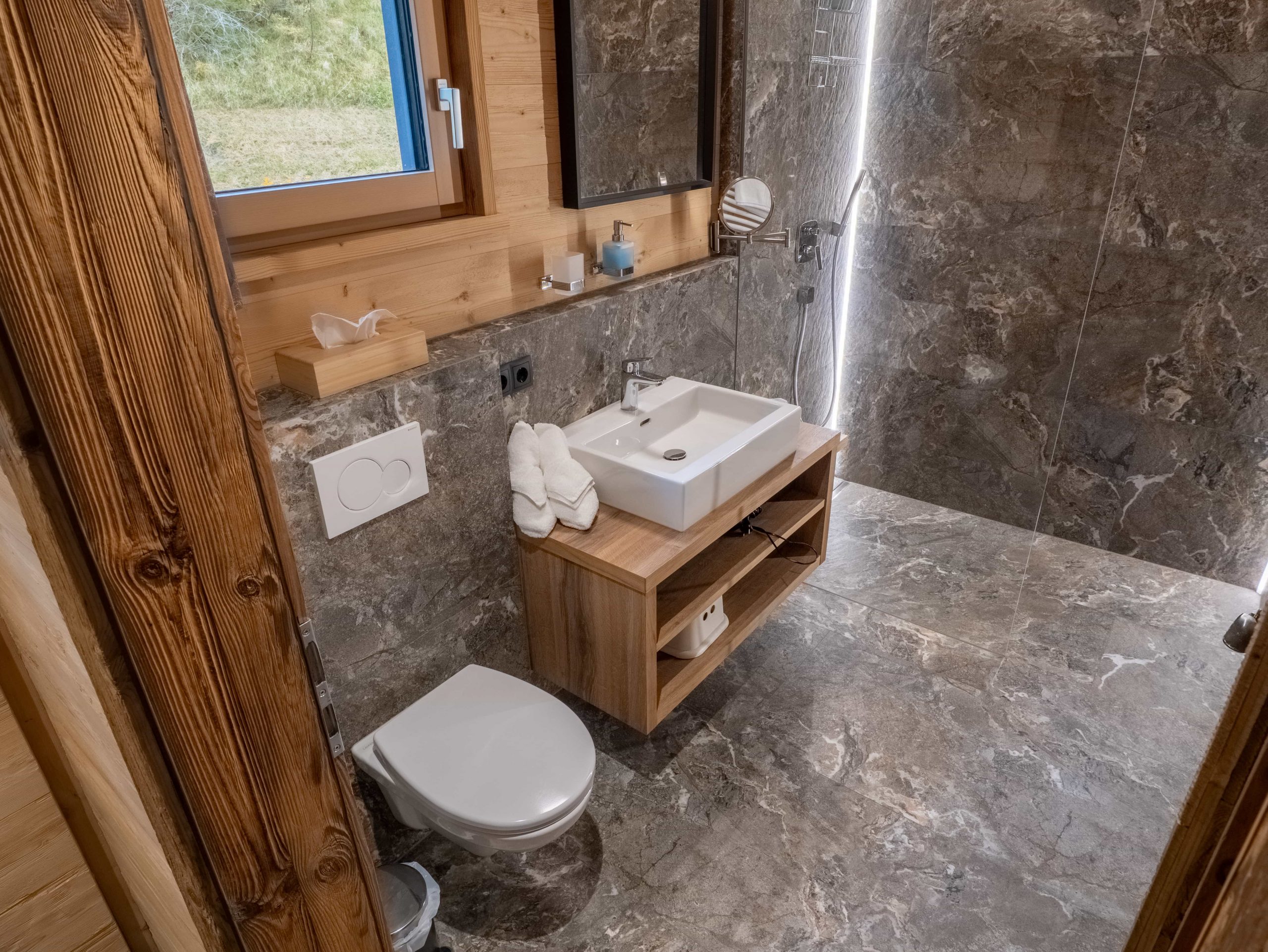 Modernes Badezimmer mit Steinfliesen an den Wänden und auf dem Boden, einer an der Wand befestigten Toilette, einem rechteckigen Waschbecken auf einem hölzernen Waschtisch, Handtüchern, einem Spiegel und einem Fenster mit Blick nach draußen ins Grüne.