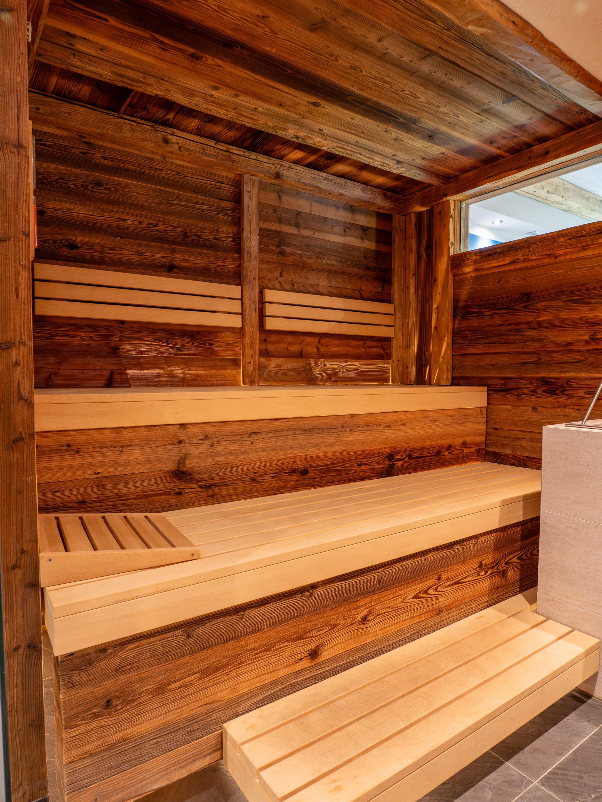 Eine moderne Innensauna mit Holzbänken und -wänden in warmen Holztönen und einem kleinen Fenster, durch das Tageslicht in den Raum fällt.
