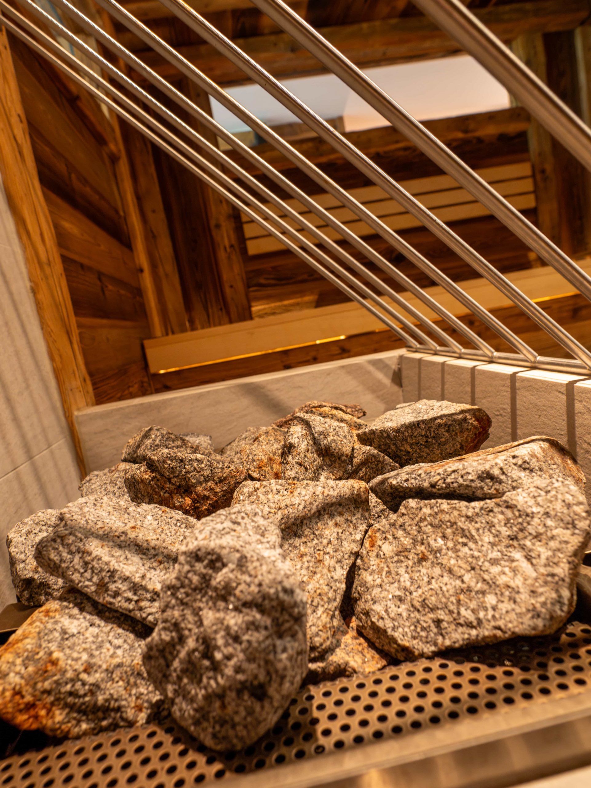 Eine Nahaufnahme von Saunasteinen, die auf einem Metallgitter gestapelt sind, mit Holzwänden und schrägen Metallgeländern im Hintergrund. Die Perspektive ist nach oben geneigt, was die rustikale Umgebung der Sauna unterstreicht.
