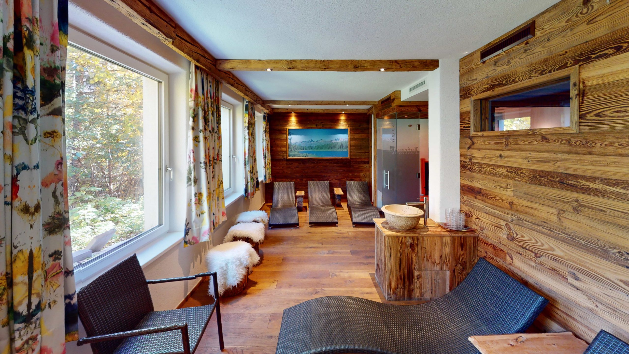 Ein gemütlicher Wellness-Raum mit Holzwänden und -böden, Lounge-Sesseln aus Korbgeflecht, flauschigen Hockern und großen Fenstern mit geblümten Vorhängen, die natürliches Licht hereinlassen. Ein Gemälde und eine Deko-Schale stehen auf einem Holzsims.
