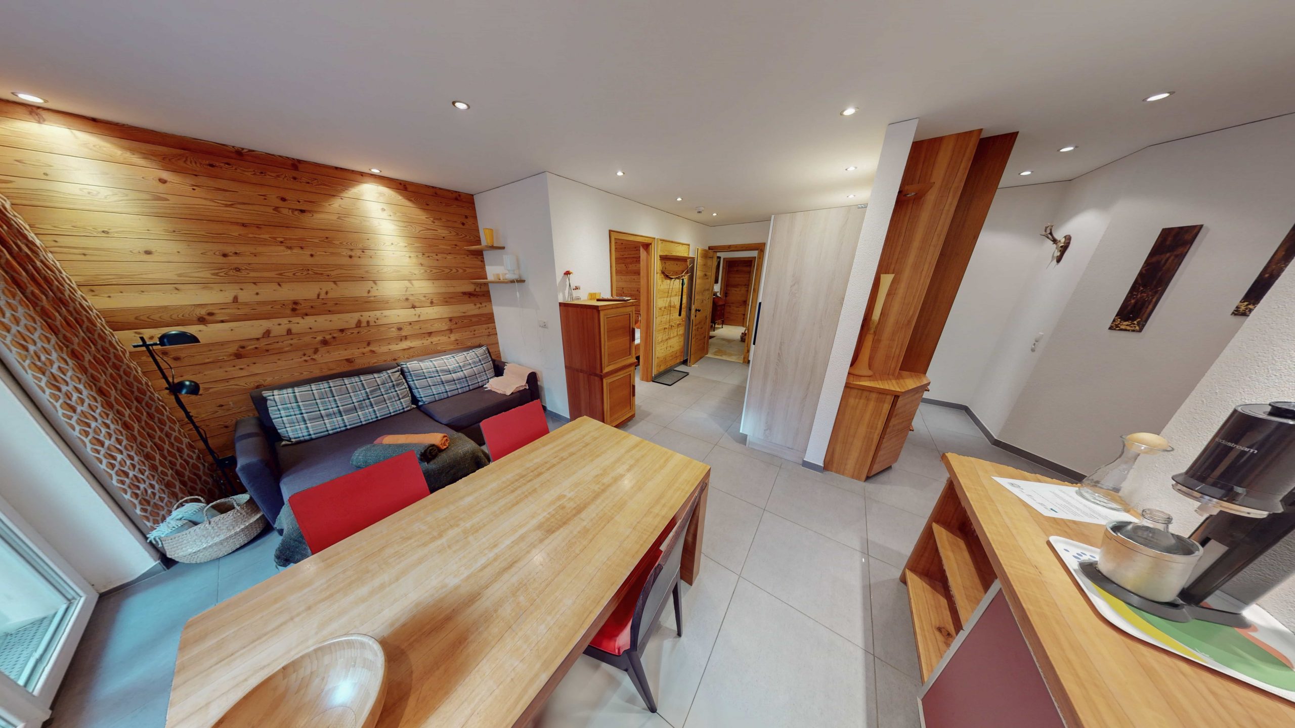 Ein gemütlicher, moderner, offener Wohnbereich mit Holzakzenten, einem Esstisch, einem Sofa und Küchengeräten. Helle Wände und geflieste Böden schaffen eine helle, einladende Atmosphäre.