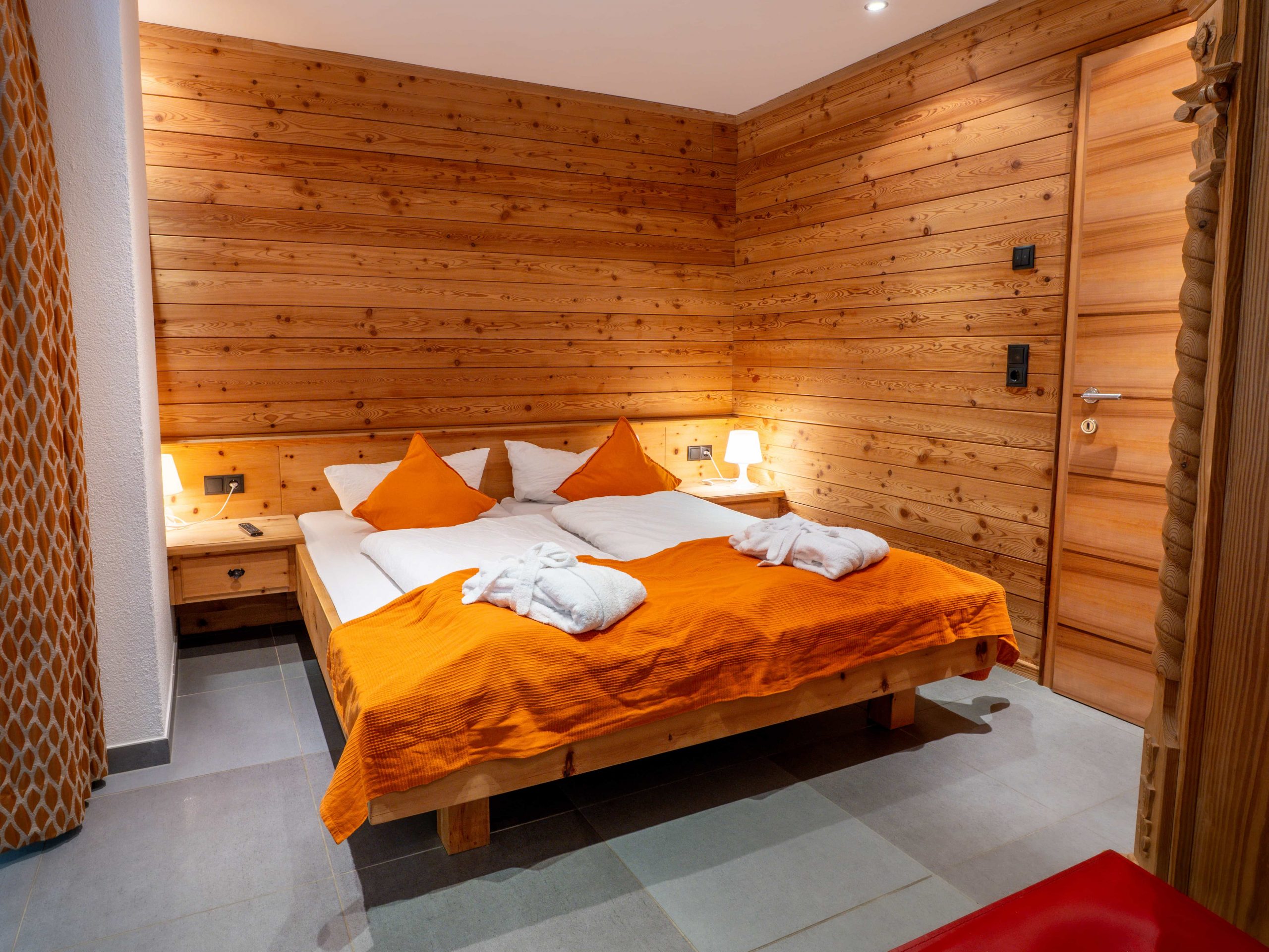 Ein gemütliches Schlafzimmer mit Holzwänden, einem Doppelbett mit weißer Bettwäsche, orangefarbenen Kissen und einer Decke, zwei Nachttischlampen und gefalteten weißen Bademänteln auf dem Bett. Das Zimmer hat eine warme und einladende Atmosphäre.