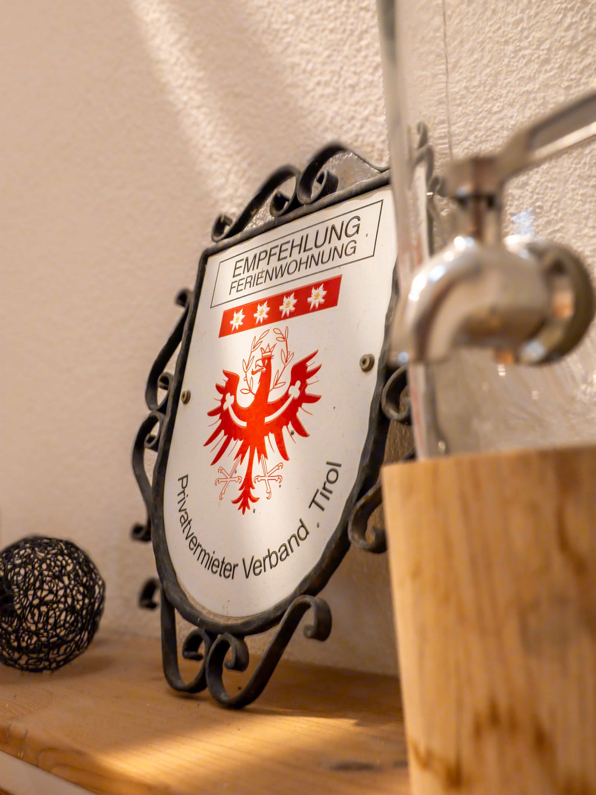 Eine dekorative Metallplakette mit einem roten Adler und Sternen, auf der "Empfehlung" steht. Ferienwohnung. Privatvermieter Verband Tirol, ruht auf einem Holzregal an der Wand, neben einem Glasbehälter und einer schwarzen Zierkugel.