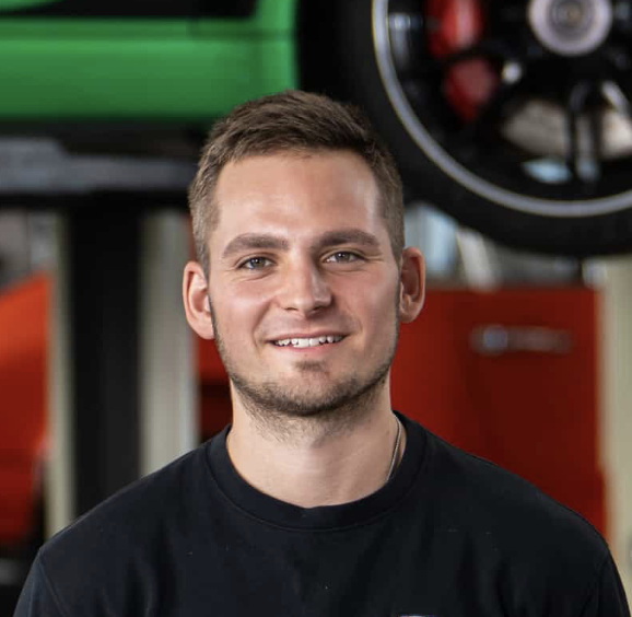 Ein lächelnder Mann mit kurzen braunen Haaren und gestutztem Bart trägt ein schwarzes Hemd und steht in einem Innenraum vor einer Autoausrüstung und einem angehobenen Fahrzeugreifen im Hintergrund.