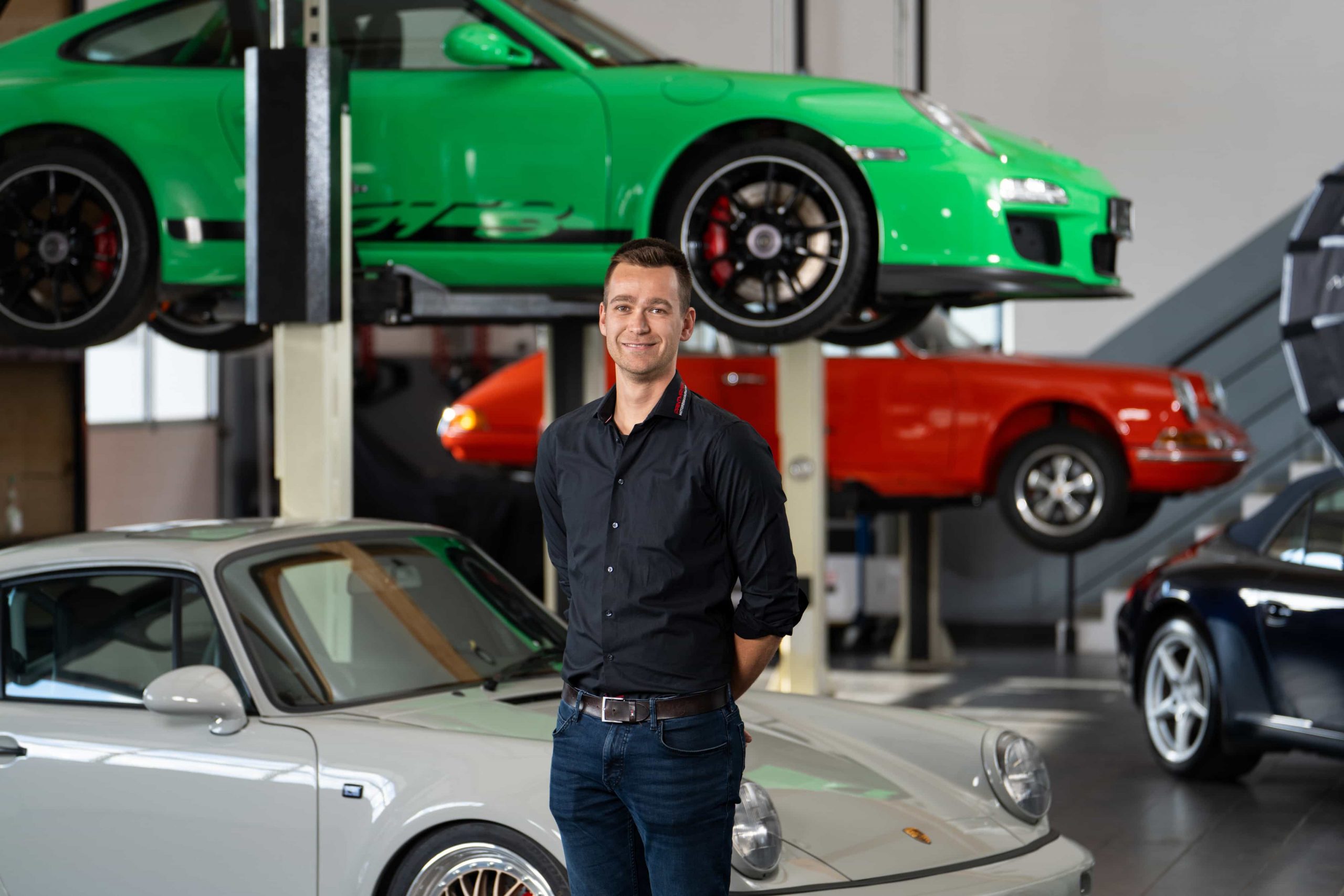 paintmayer_portrait_christian_paintmayer_oktober2025_8©invisions Ein Mann in einem schwarzen Hemd steht lächelnd vor einem silbernen Porsche, während zwei andere Porsche - ein grüner und ein roter - auf Hebebühnen im Hintergrund in einer Garage hochgefahren werden.