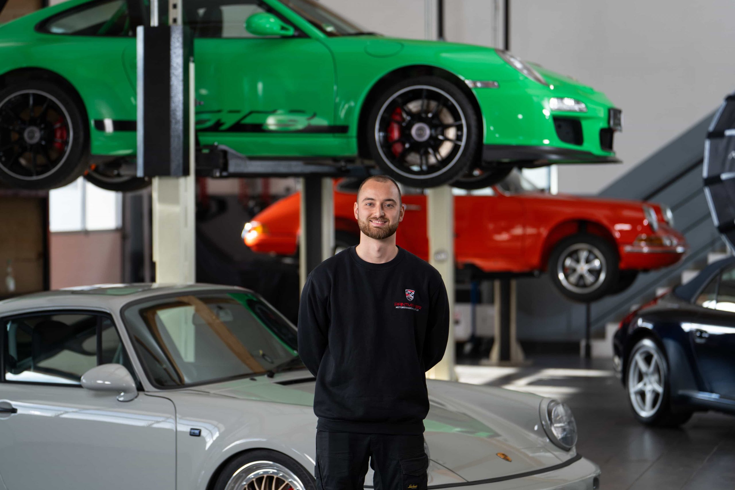paintmayer_portrait_jonas_oktober2025_6©invisions Ein Mann steht lächelnd in einer Garage, umgeben von mehreren Porsche-Autos, darunter ein grüner auf einer Hebebühne über ihm und andere, die um ihn herum geparkt sind. Die Umgebung scheint eine Autowerkstatt oder ein Ausstellungsraum zu sein.