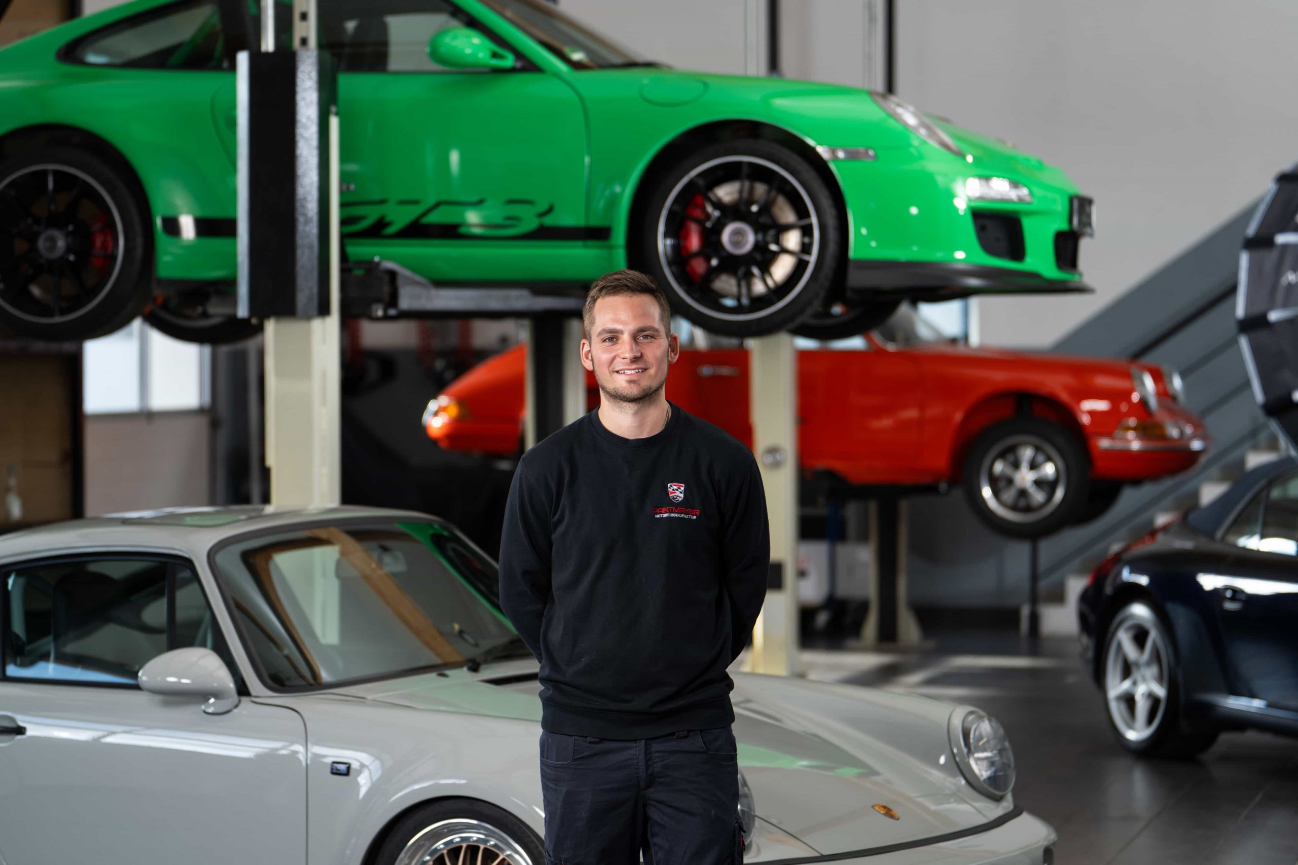 paintmayer_portrait_justin_oktober2025_1©invisions Ein Mann in einem schwarzen Sweatshirt steht lächelnd in einer Garage mit mehreren klassischen Sportwagen, darunter ein grünes Auto auf einer Hebebühne und ein silbernes Auto vor ihm.