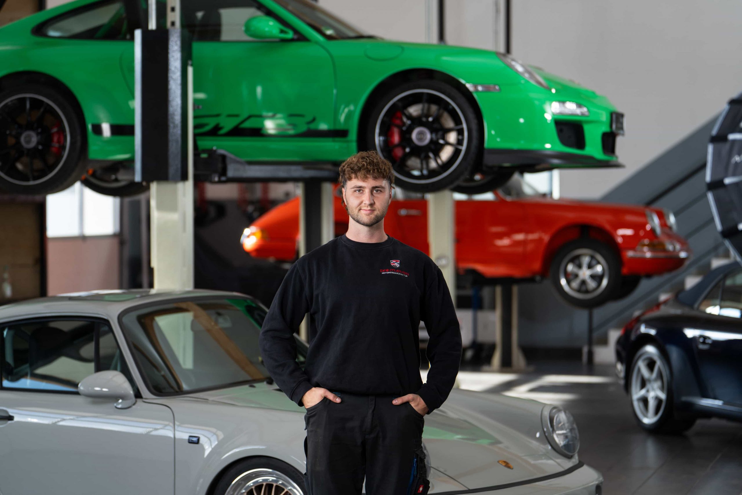 paintmayer_portrait_markus_oktober2025_4©invisions Ein Mann in einem schwarzen langärmeligen Hemd steht in einer Garage mit mehreren Sportwagen, darunter ein grünes Auto auf einer Hebebühne, ein silbernes Auto davor und ein rotes Auto im Hintergrund.