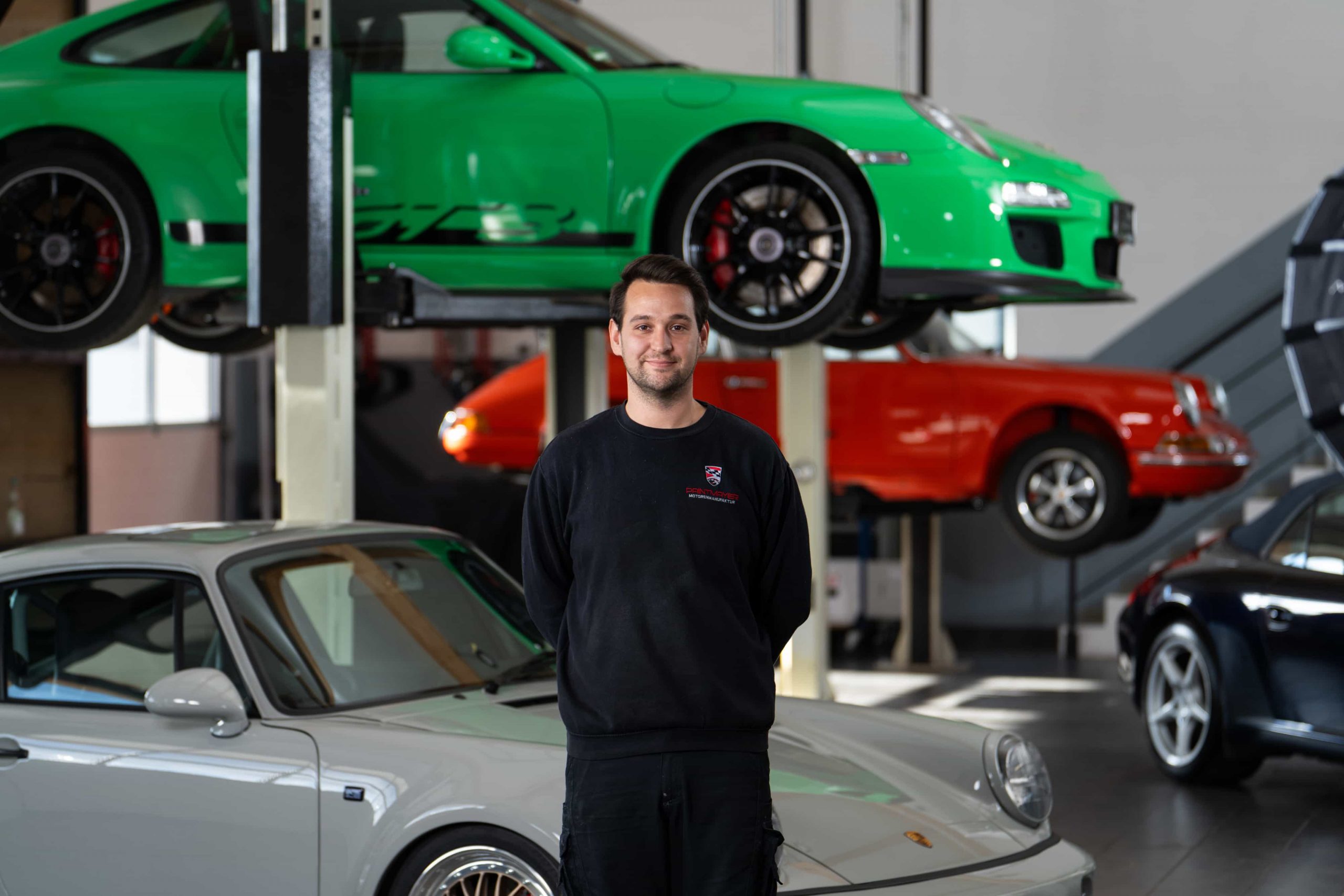 paintmayer_portrait_michael_oktober2025_2©invisions Ein Mann in einem schwarzen Sweatshirt steht in einer Garage mit klassischen und modernen Porsche-Fahrzeugen, darunter ein grünes Auto auf einer Hebebühne hinter ihm und ein silbernes Auto im Vordergrund.