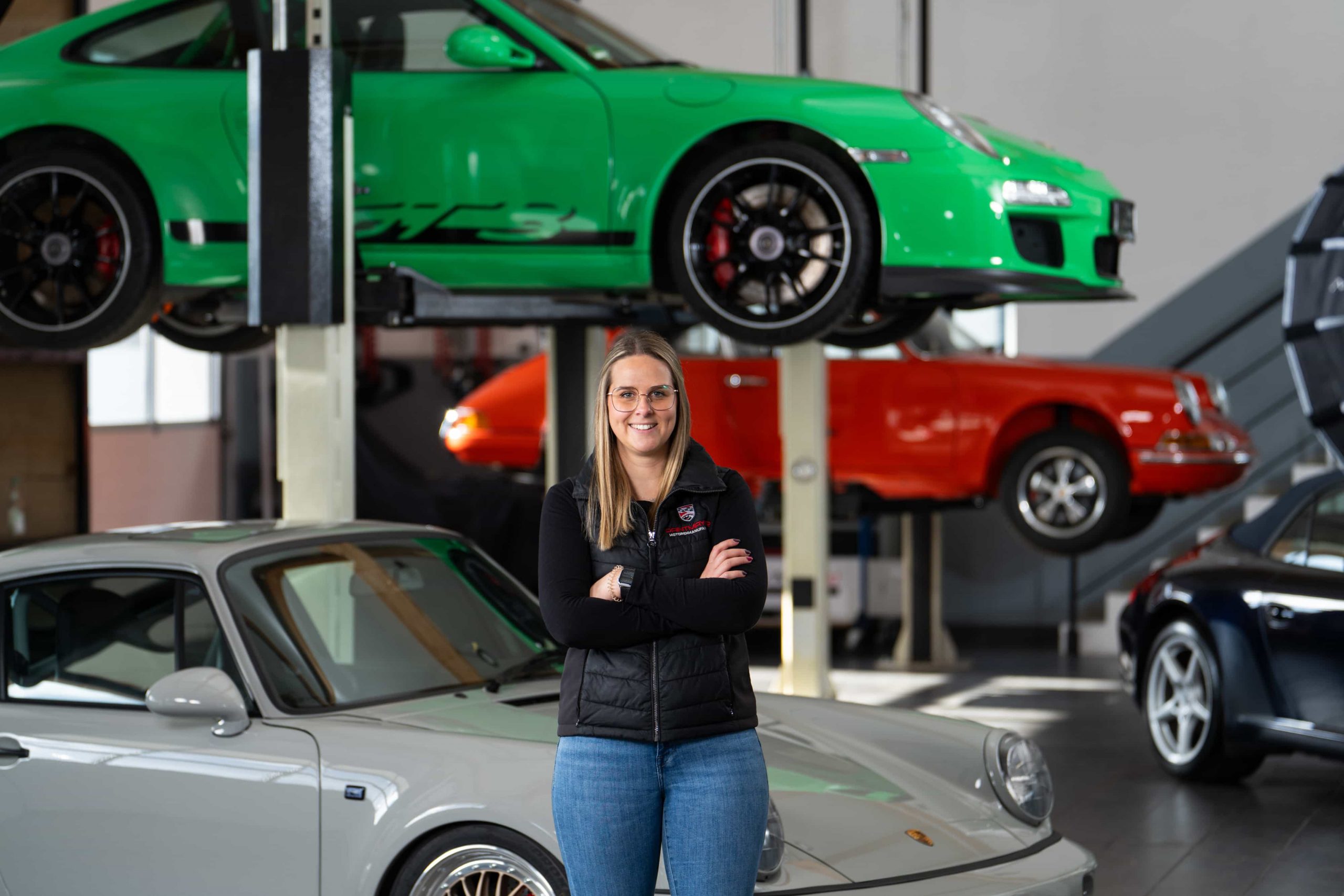 paintmayer_portrait_tamara_zwick_oktober2025_7©invisions Eine Frau mit langen blonden Haaren, Brille und schwarzer Weste steht lächelnd mit verschränkten Armen in einer Garage, umgeben von mehreren klassischen und modernen Porsche-Fahrzeugen, darunter ein grüner Wagen, der im Hintergrund angehoben wird.