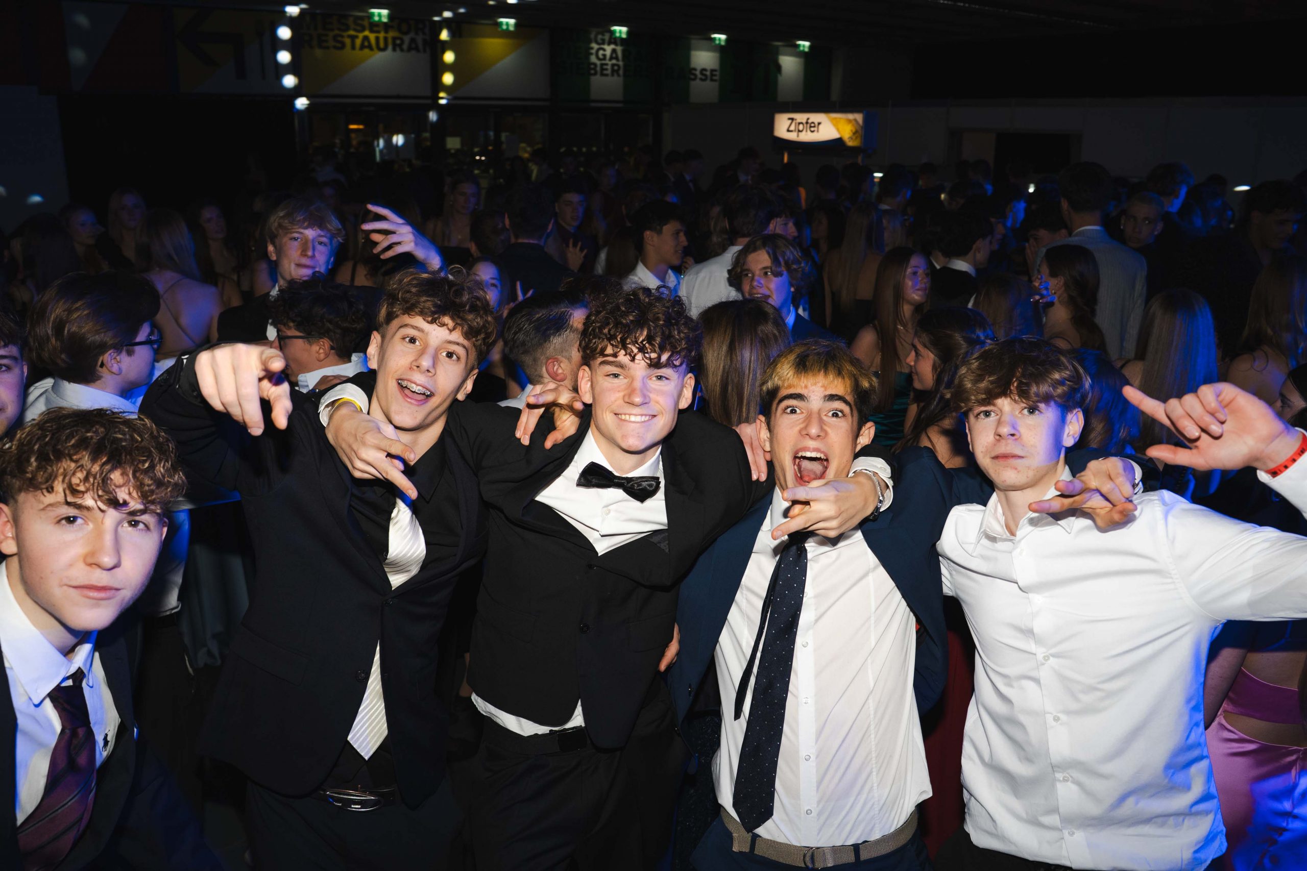 Eine Gruppe von fünf jungen Männern in formeller Kleidung lächelt und posiert enthusiastisch für die Kamera auf einer überfüllten Party oder Veranstaltung in einer Halle, mit vielen Menschen im Hintergrund unter bunten Lichtern.